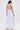 A woman stands facing away in the TOO COOL HIGH LOW TOP, a sleeveless white halter-style top with ruffles and a ruffled hem. She holds a white woven clutch, wears large earrings, has her hair in a low ponytail, against a plain white background.