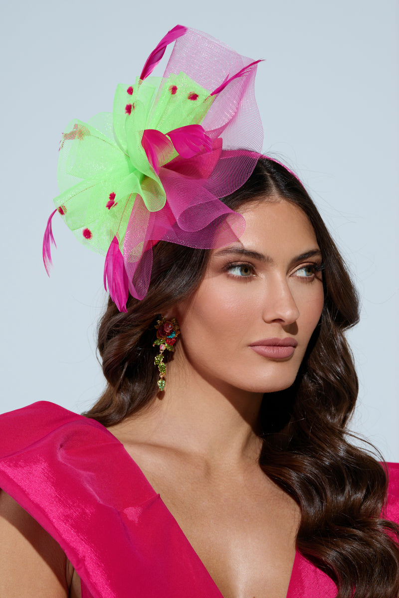 A woman with long wavy hair wears a vibrant pink dress and the TO THE RACES FASCINATOR, showcasing pink and green tulle with feather details. Her matching earrings enhance the elaborate headpiece as she gazes off to the side against a plain backdrop.