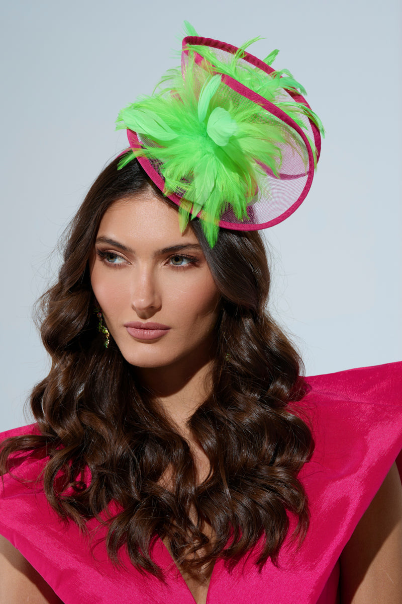 The woman, sporting long, wavy hair, wears a bright pink dress with structured shoulders and a TO THE DERB FASCINATOR in vibrant lime green. The feathered accessory adds flair against the plain backdrop as she gazes slightly to the side with a neutral expression. 0