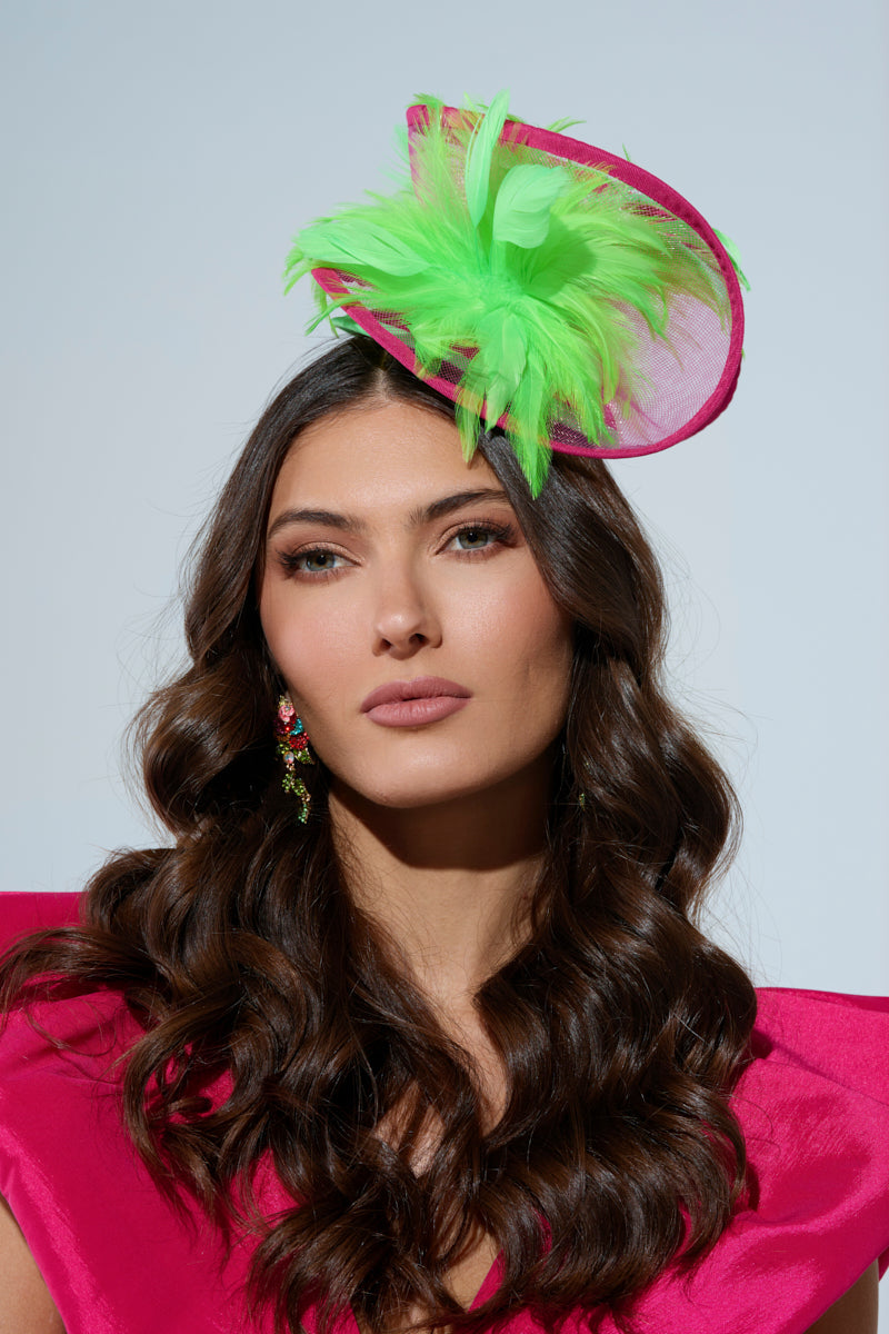 A woman with long brown hair displays a maximalist style, dressed in a bright pink outfit, wearing the "TO THE DERB FASCINATOR" adorned with green feathers and colorful earrings. She maintains a neutral expression against a plain background. 1