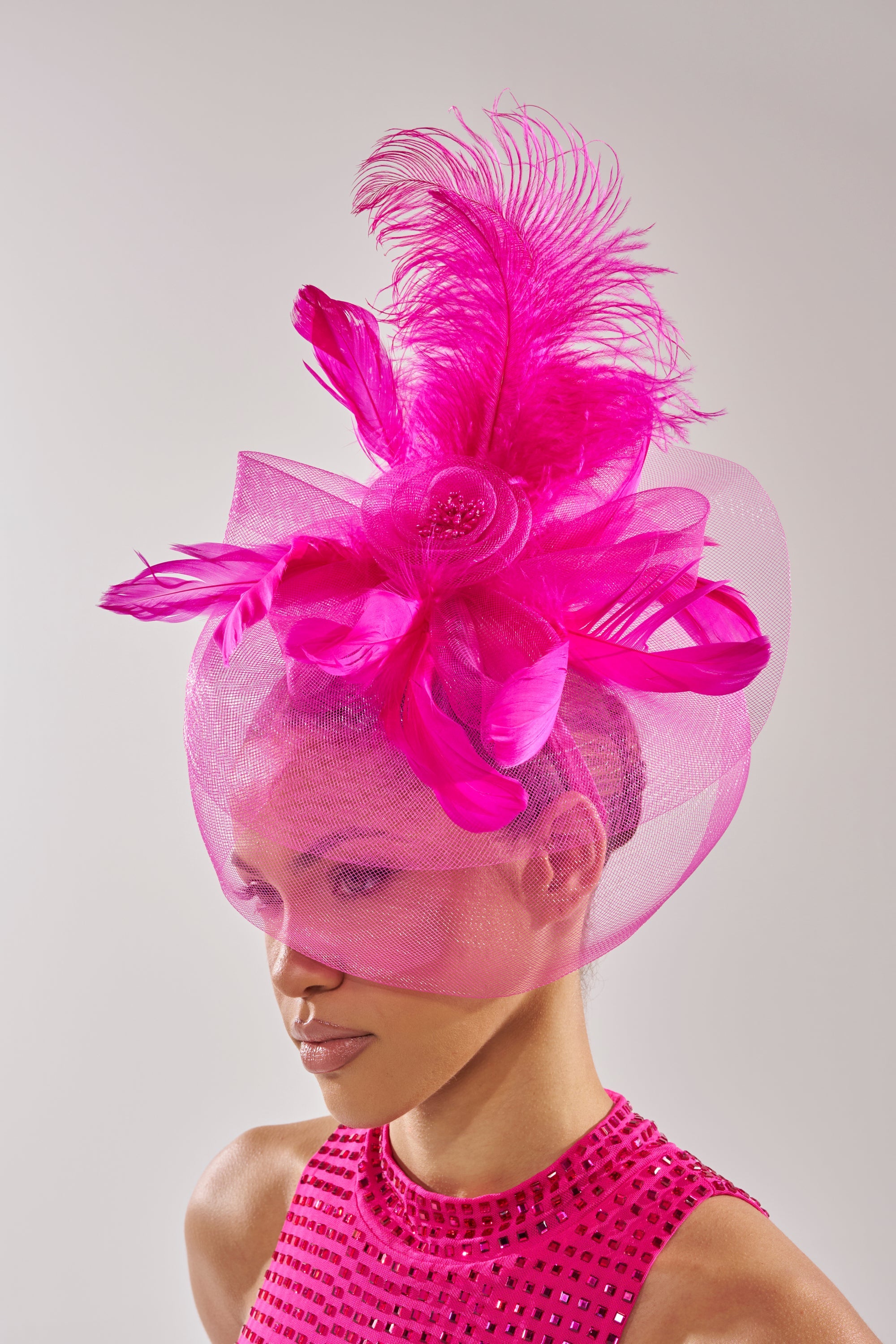 A person wears the DERBY DAY PINK FASCINATOR, featuring large pink feathers, mesh, and a satin headband that partially covers their face, paired with a sleeveless pink top embellished with rhinestones. 0