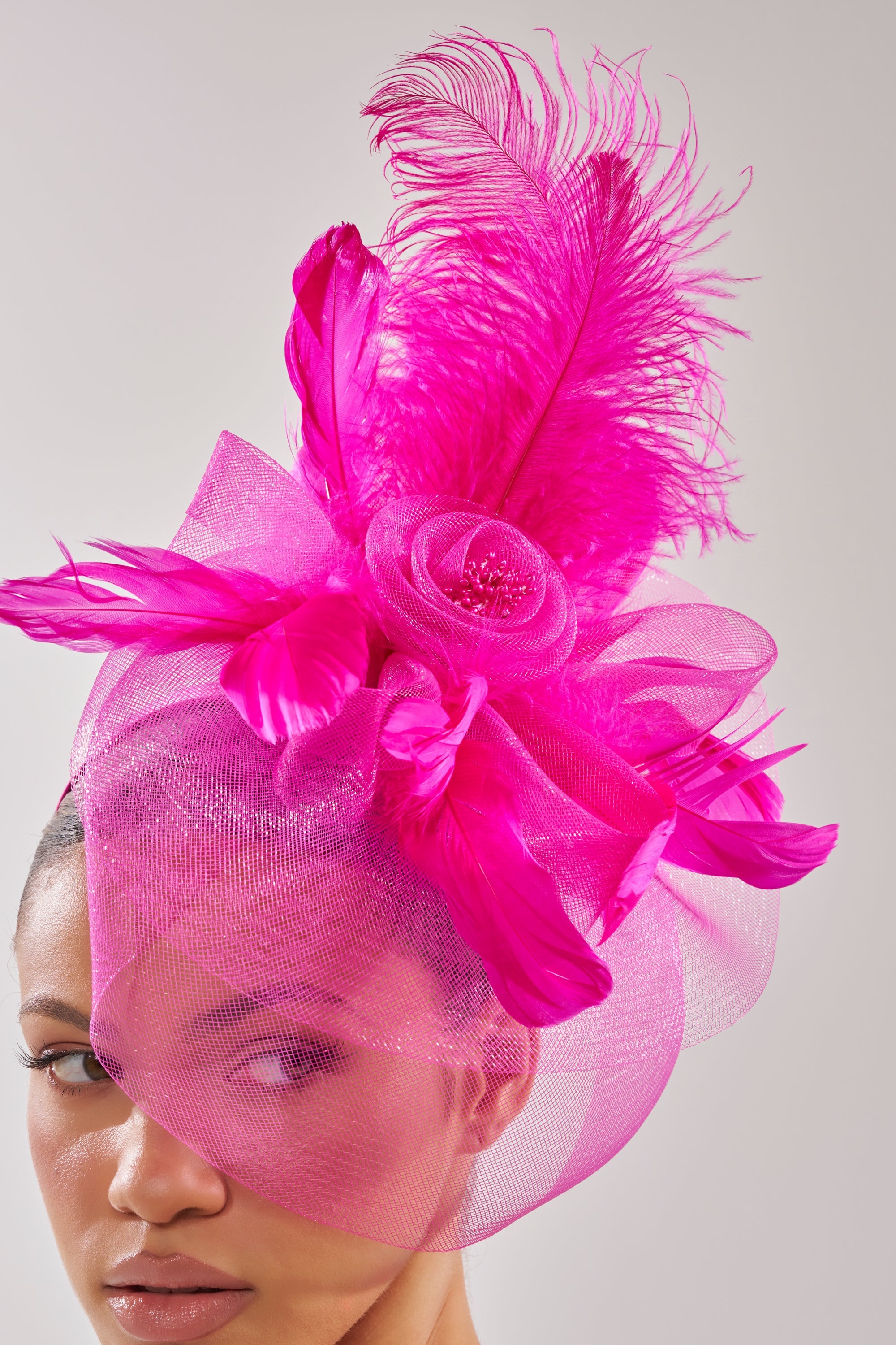 A woman wears the DERBY DAY PINK FASCINATOR with large feather accents, mesh, and ribbon on a pink satin headband. The bright pink accessory partially covers her face against a plain, light background. 1