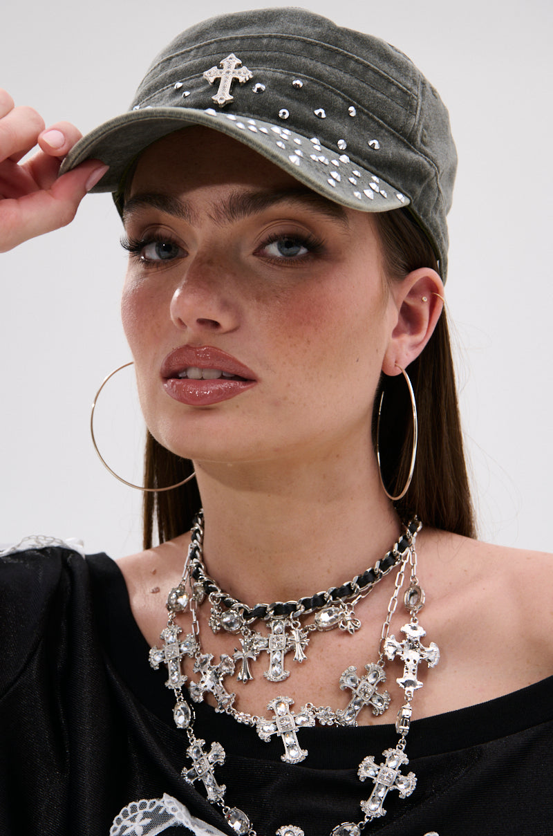 A woman wears the GET WITH IT HAT, a green military-style cap with rhinestone and cross accents. She pairs it with large hoops, glossy lipstick, layered cross necklaces, and holds the brim while looking at the camera.