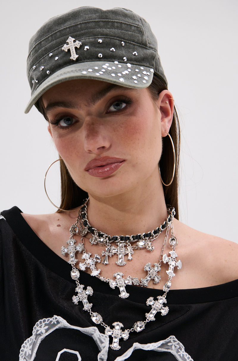 A woman wears the GET WITH IT HAT, a studded military-style cap with rhinestone embellishments and a cross emblem, paired with large hoop earrings, layered silver cross necklaces, and a black off-the-shoulder top. Her straight brown hair frames natural makeup.
