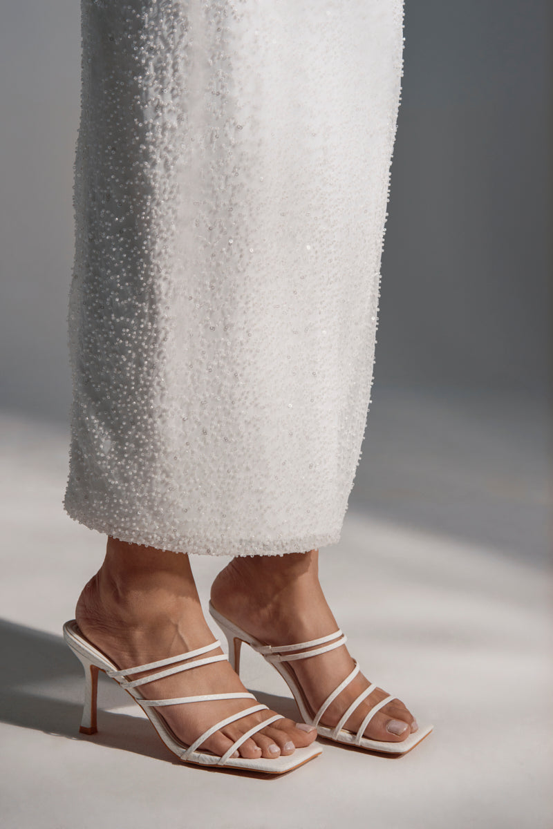 Someone stands on a light surface wearing a long, textured white skirt and the AZALEA WANG HUGHIE WHITE SNAKE SANDAL, featuring open toes and thin stiletto heels.