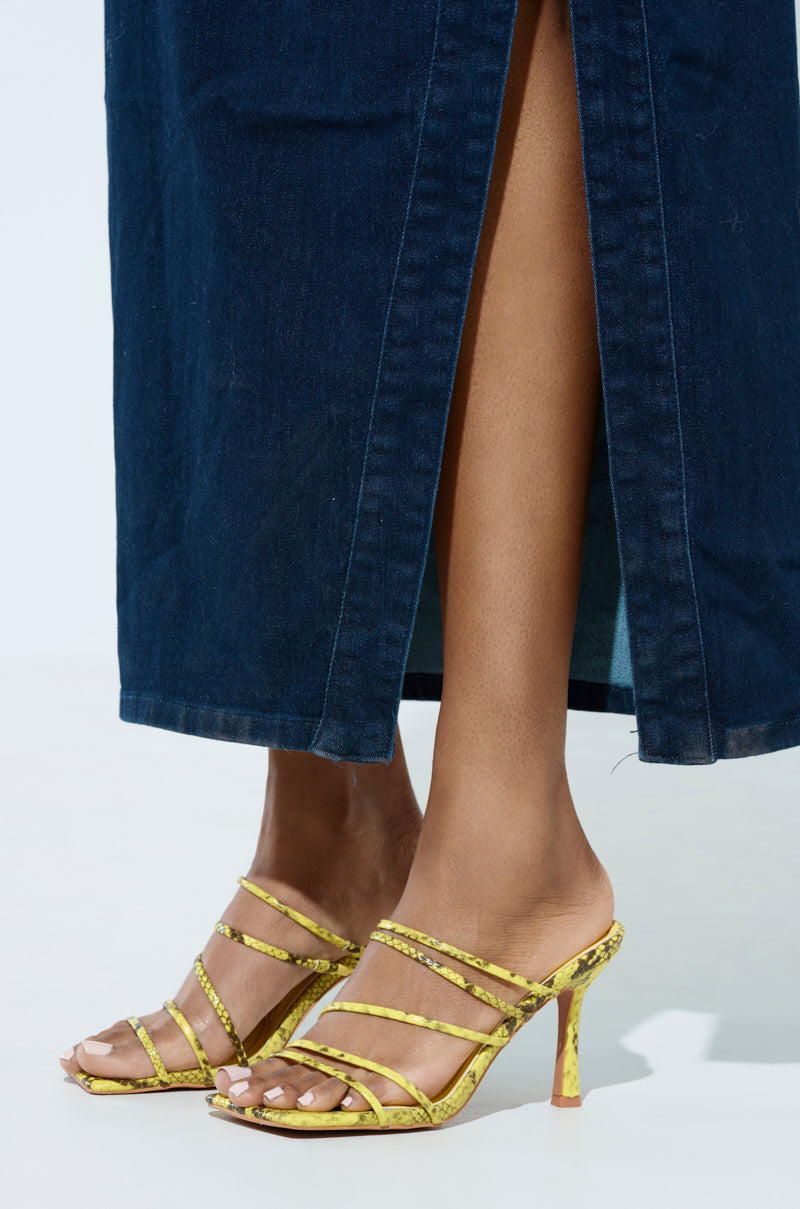 A person is standing on a light surface wearing the AZALEA WANG HUGHIE YELLOW SNAKE SANDAL, featuring sleek vegan leather and an open square toe. Their outfit includes a dark blue denim skirt with a center slit that reveals their lower legs.