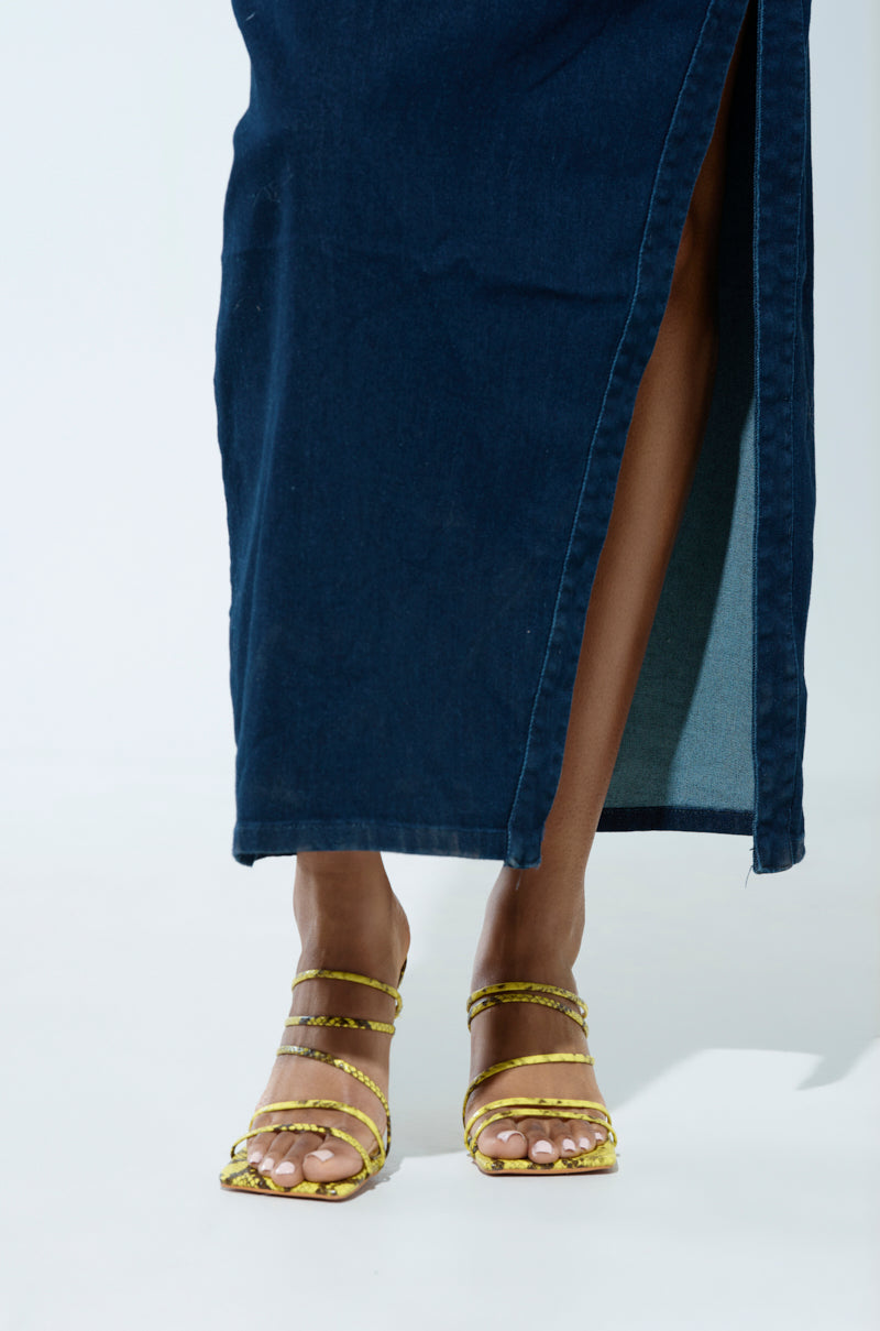 Against a light backdrop, someone wears a long dark denim skirt with a slit and open-toe AZALEA WANG HUGHIE Yellow Snake Sandals, revealing light pink toenail polish.