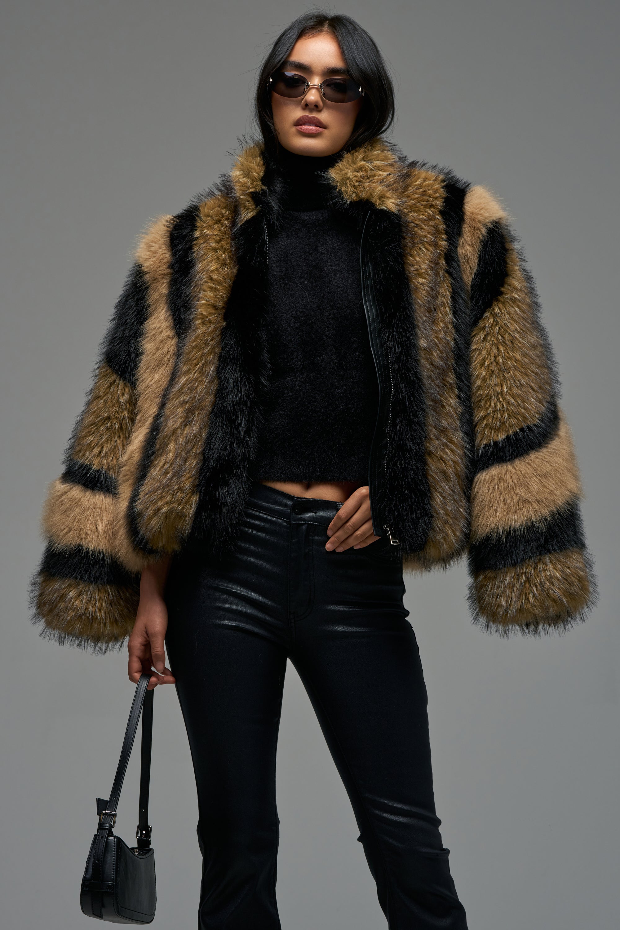 A woman stands against a plain gray background wearing sunglasses, a black turtleneck, black leather pants, and holding a small black handbag, styled with the HURRICANE PATCHWORK FAUX FUR jacket.