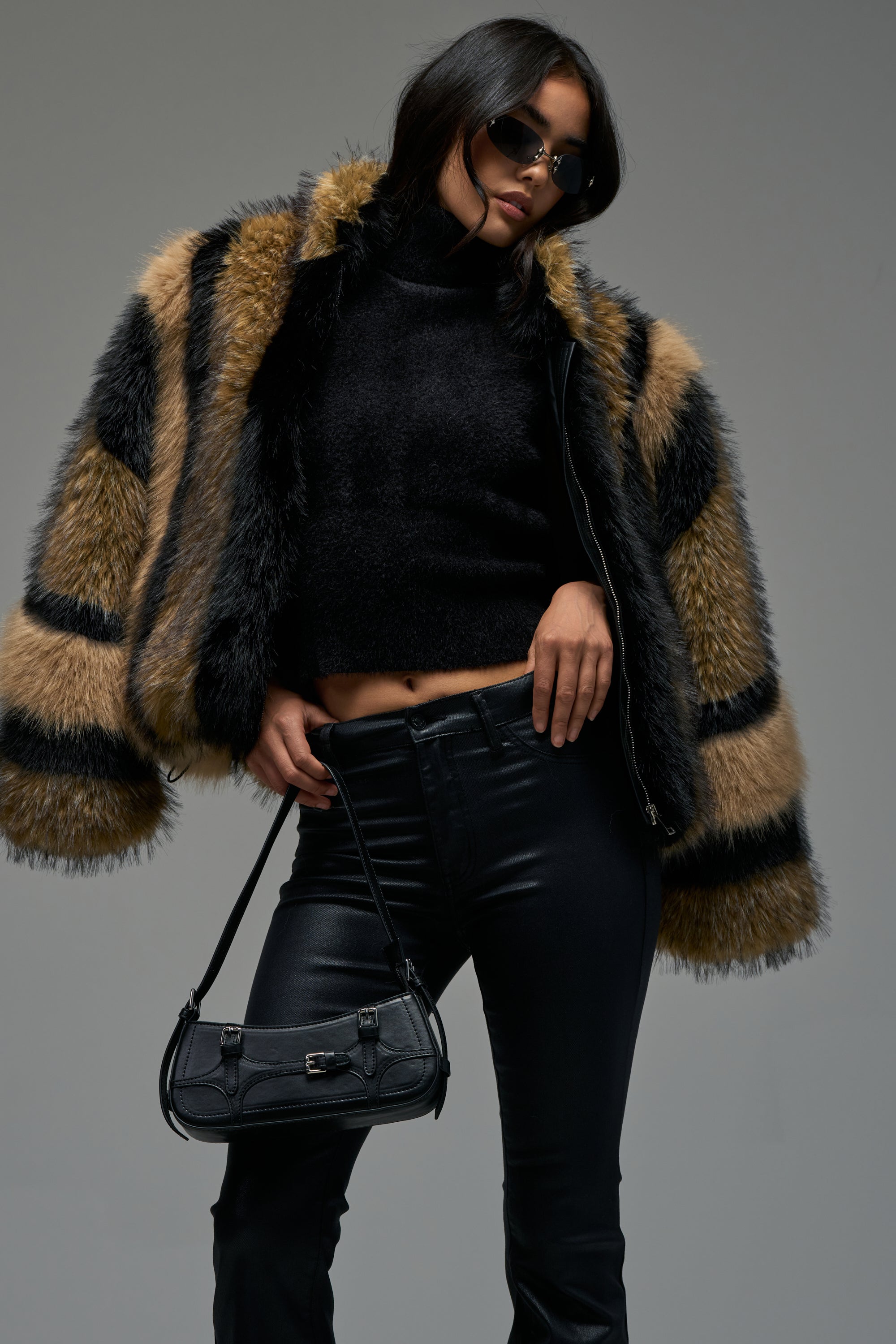 A woman stands confidently against a plain background, wearing the HURRICANE PATCHWORK FAUX FUR jacket in black and tan stripes, paired with a black turtleneck, leather pants, sunglasses, and a small black handbag.