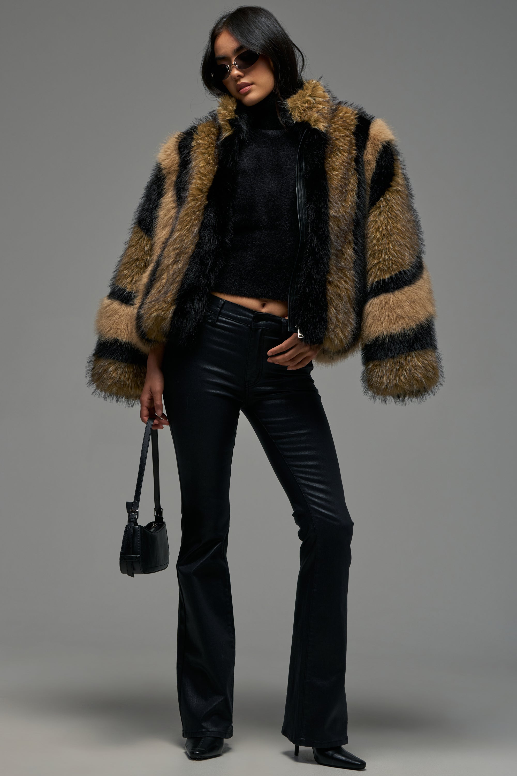 Wearing the HURRICANE PATCHWORK FAUX FUR jacket, a woman in sunglasses makes a bold winter statement by pairing it with a black turtleneck, black leather pants, and black boots. She carries a small black handbag while posing against a plain gray background.