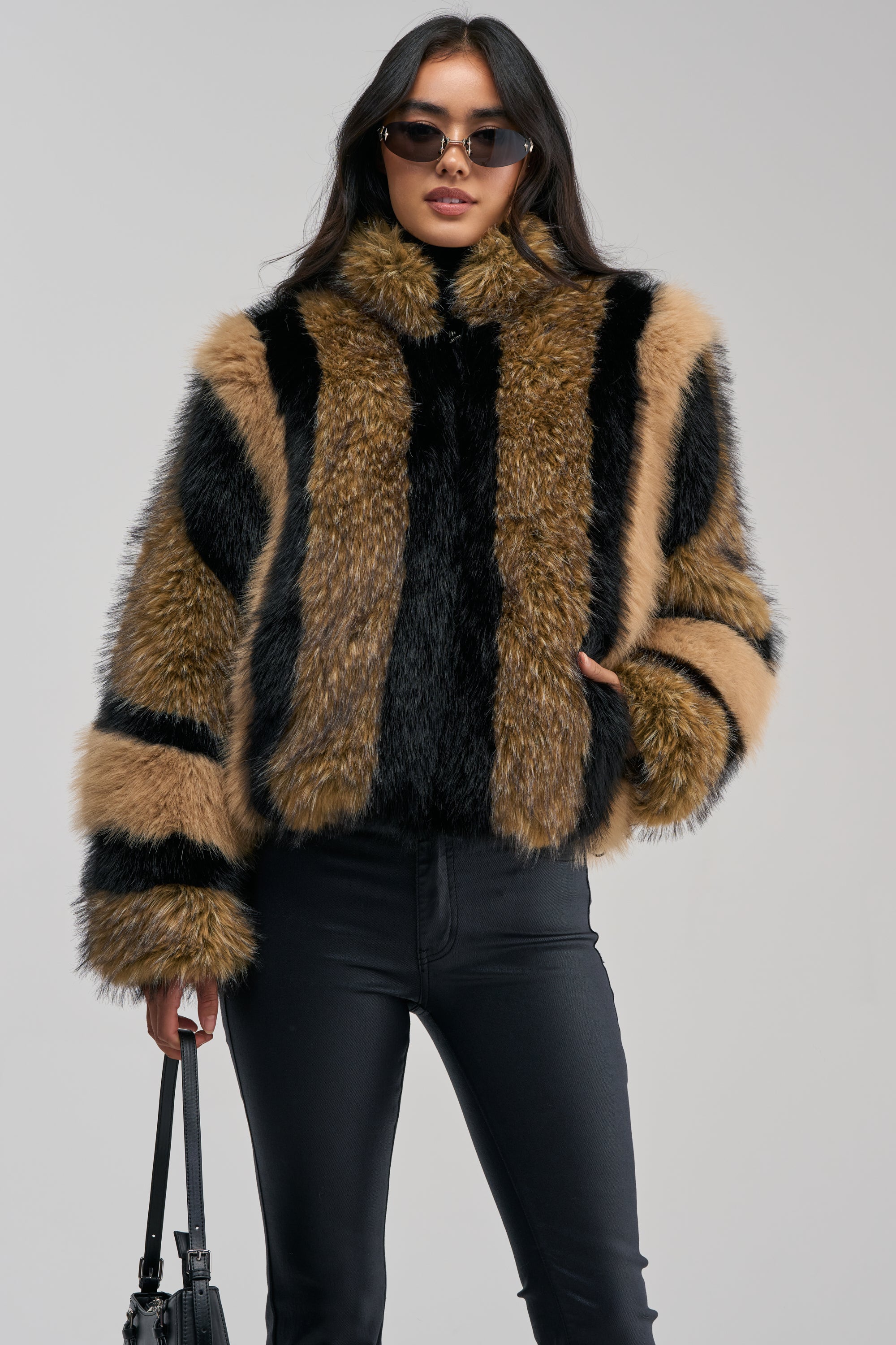 A woman stands against a plain, light-colored background, making a chic winter statement in the HURRICANE PATCHWORK FAUX FUR jacket, paired with sunglasses, black pants, and a black handbag.