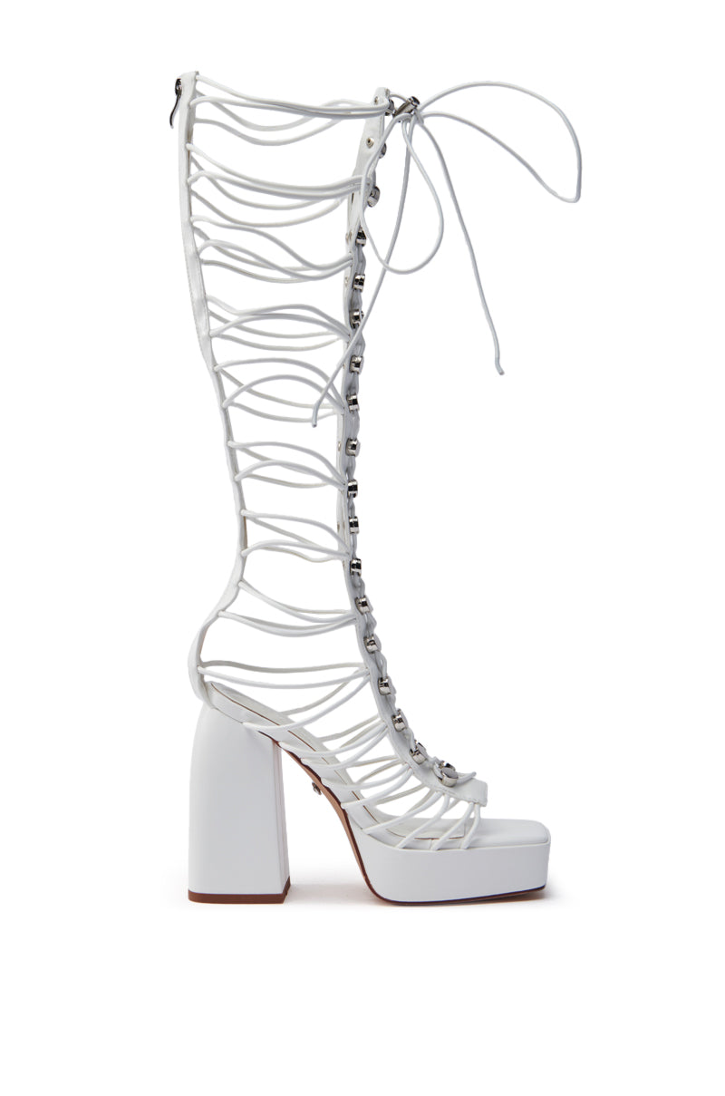 The AZALEA WANG HUYEN WHITE GLADIATOR SANDAL is a high-heeled sandal with a chunky heel and thick platform sole, made from vegan leather. It has multiple thin straps, silver-tone studs, and a knee-high lace-up closure.