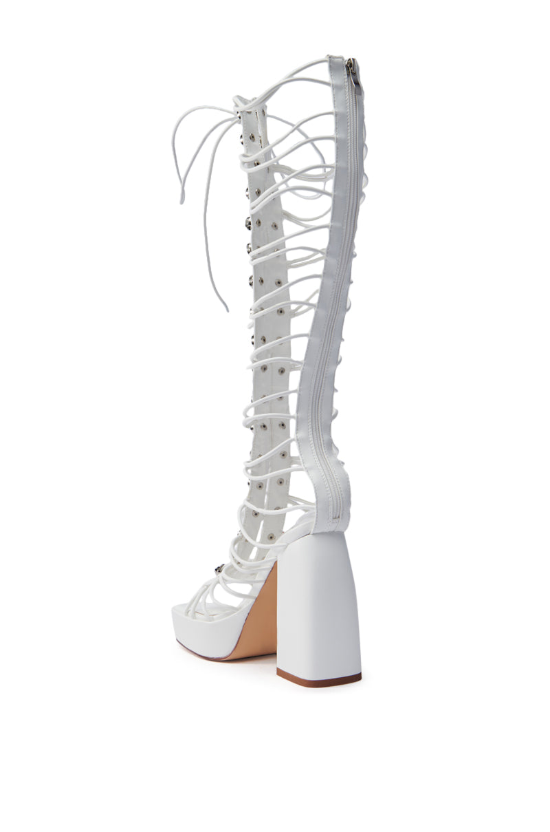 The AZALEA WANG HUYEN WHITE GLADIATOR SANDAL is a high-heeled, open-toed sandal with a gladiator lace-up design. It features a chunky platform, multiple studded straps in vegan leather, and a back zipper closure. The sole is light brown.