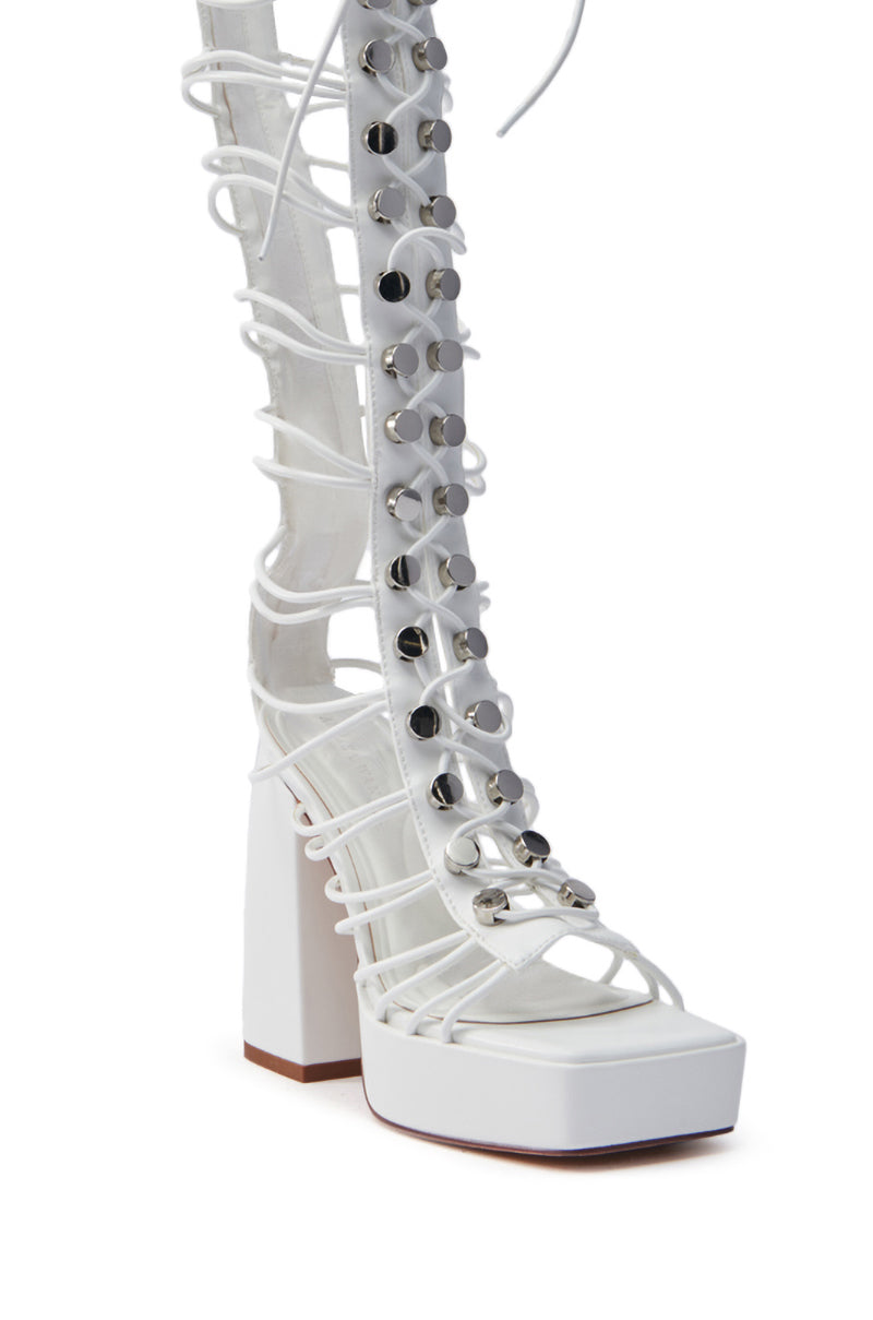 The AZALEA WANG HUYEN WHITE GLADIATOR SANDAL is a white high-heeled gladiator sandal with a platform base, made from vegan leather. It has intricate straps with silver studs up to mid-calf and a chunky block heel for style and comfort.