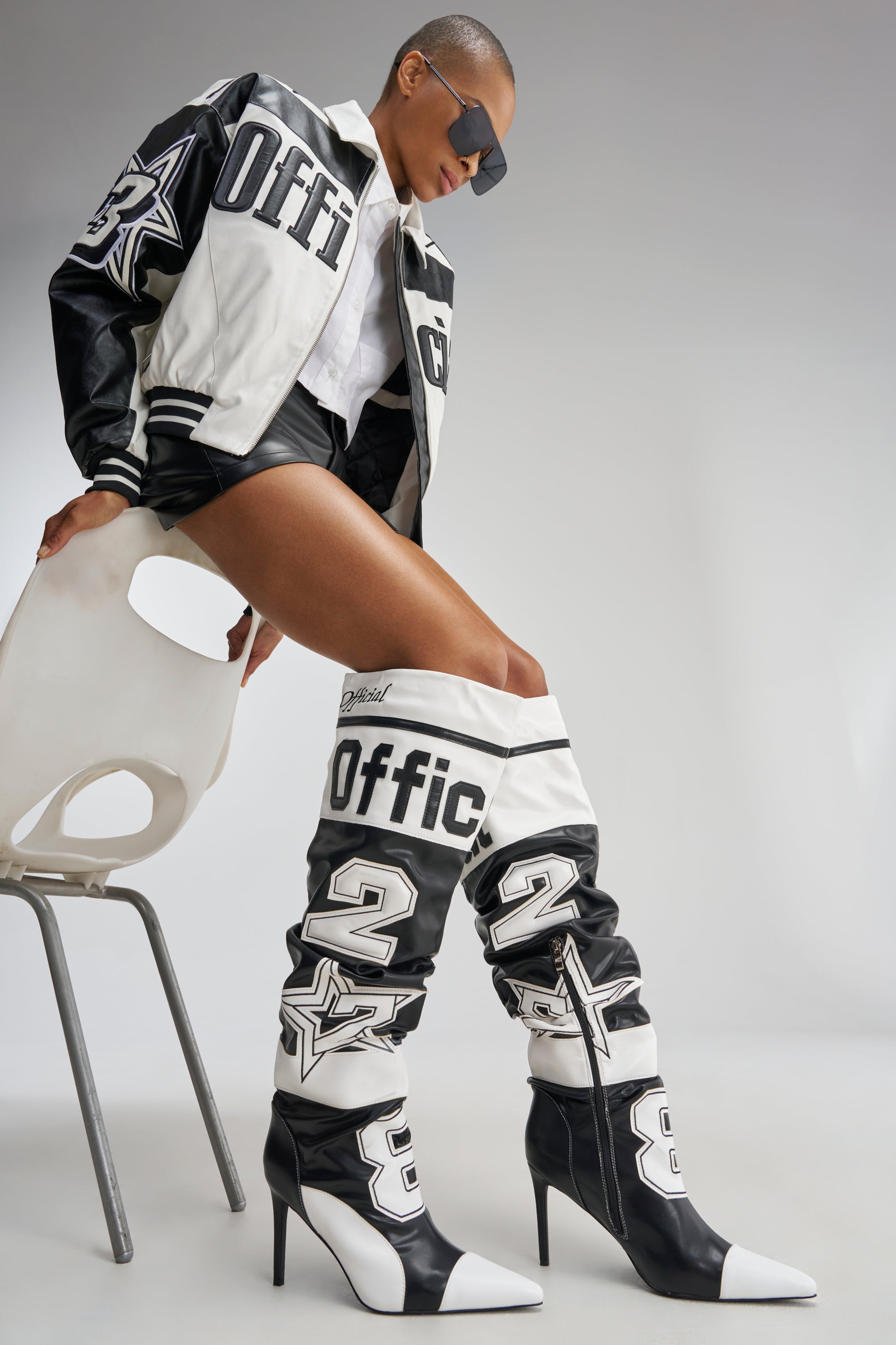 A person wearing the AKIRA GOLD HYPE COLOR BLOCKED SPORTY STILETO BOOT, styled with a black and white varsity jacket, shorts, and oversized sunglasses, sits sideways on a white chair.