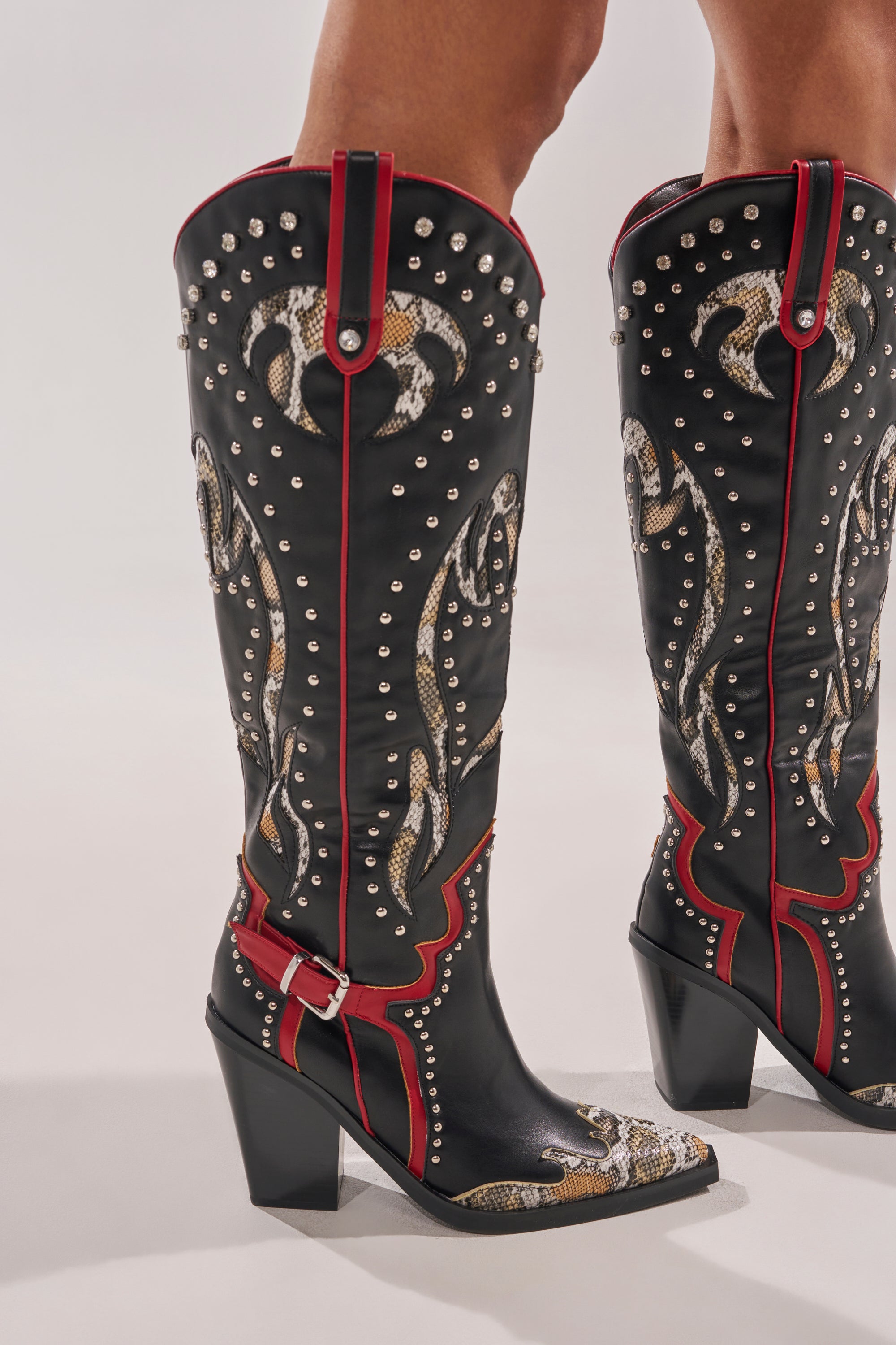Wearing the AZALEA WANG HYPERION BLACK MULTI COLOR WESTERN STUD BOOT—black knee-high boots with red trim, silver studs, snakeskin-patterned accents, and chunky high heels—this person stands on a light background. 2