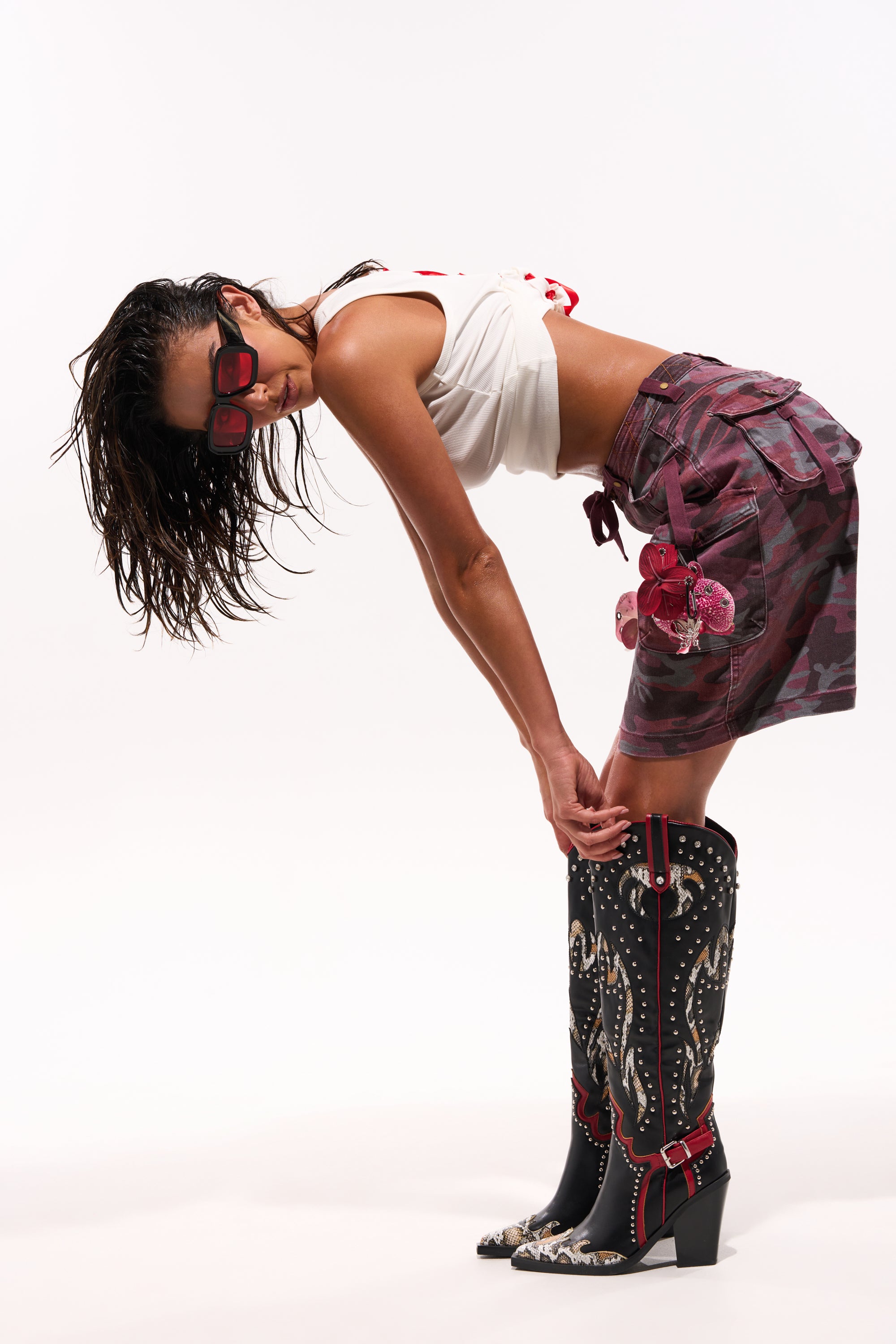 A woman with wet hair in red sunglasses, a white crop top, a purple camo skirt with butterfly patches, and AZALEA WANG HYPERION BLACK MULTI COLOR WESTERN STUD BOOTS with white stitching bends forward against a white background. 1