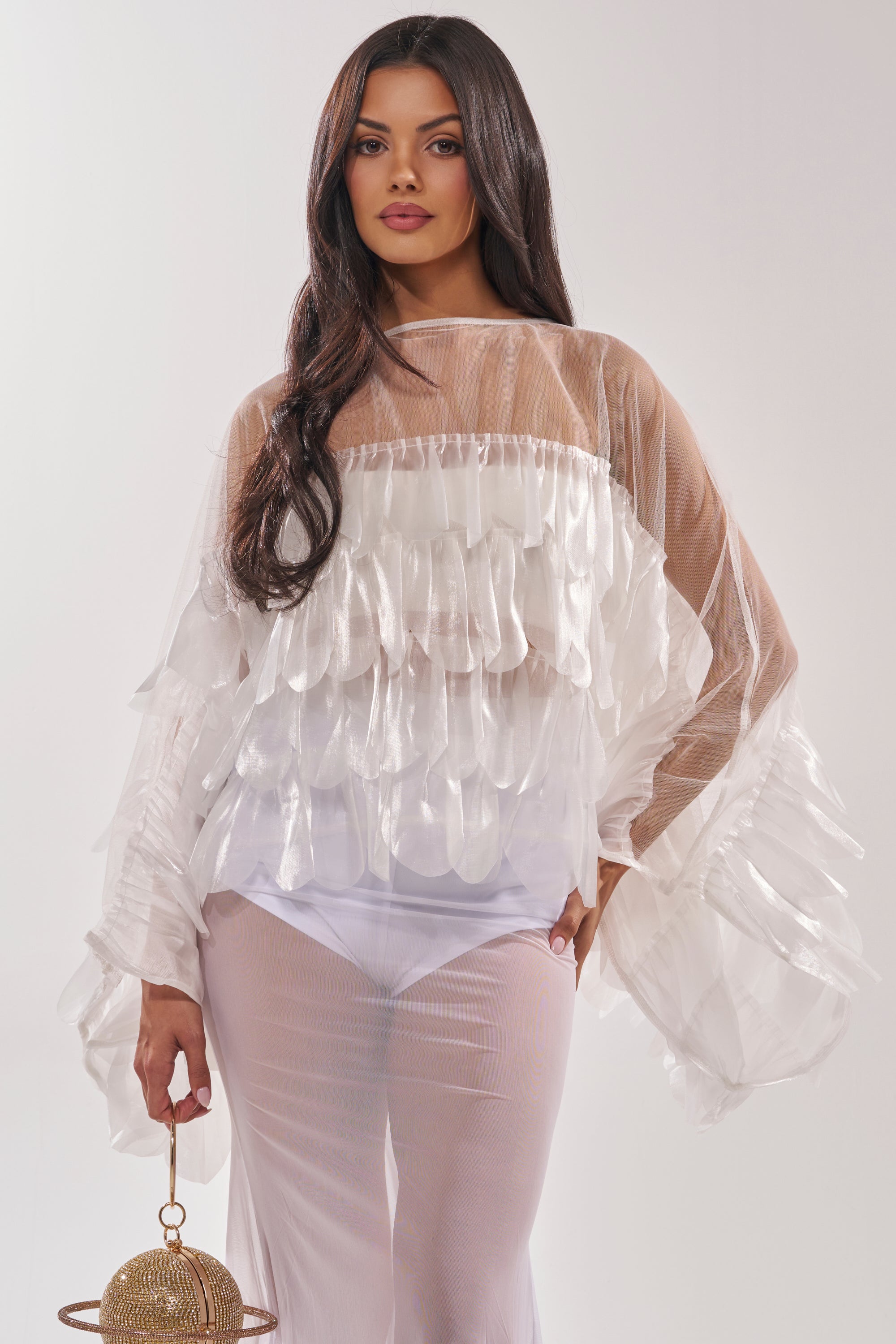 A woman with long dark hair models the RUFFLE CITY BLOUSE, a sheer mesh top with ruffled details. She pairs it with matching sheer pants and holds a small round gold purse, posing confidently against a plain light background. 1