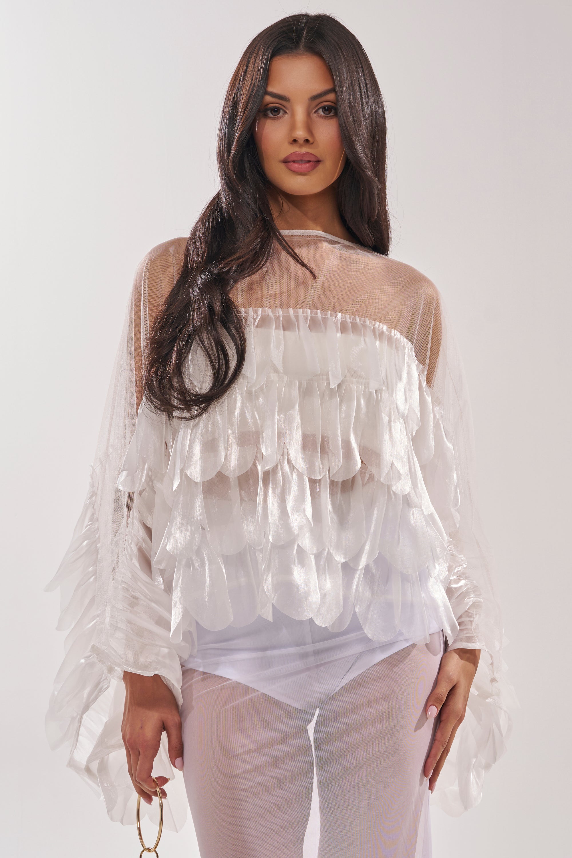 A woman with long dark hair models the RUFFLE CITY BLOUSE, which features sheer mesh panels and petal-inspired ruffle details, paired with white pants. She stands against a light background, holding a gold accessory. 2