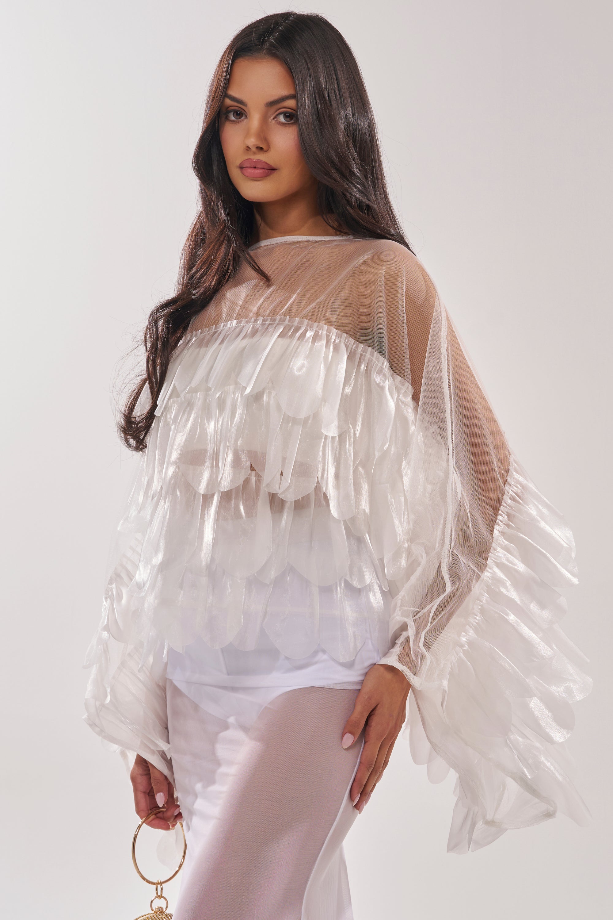 A woman with long dark hair models the RUFFLE CITY BLOUSE, which features sheer mesh panels and ruffled, feathery textures. She stands against a light background in a white outfit, holding a gold ring-shaped accessory in one hand. 3