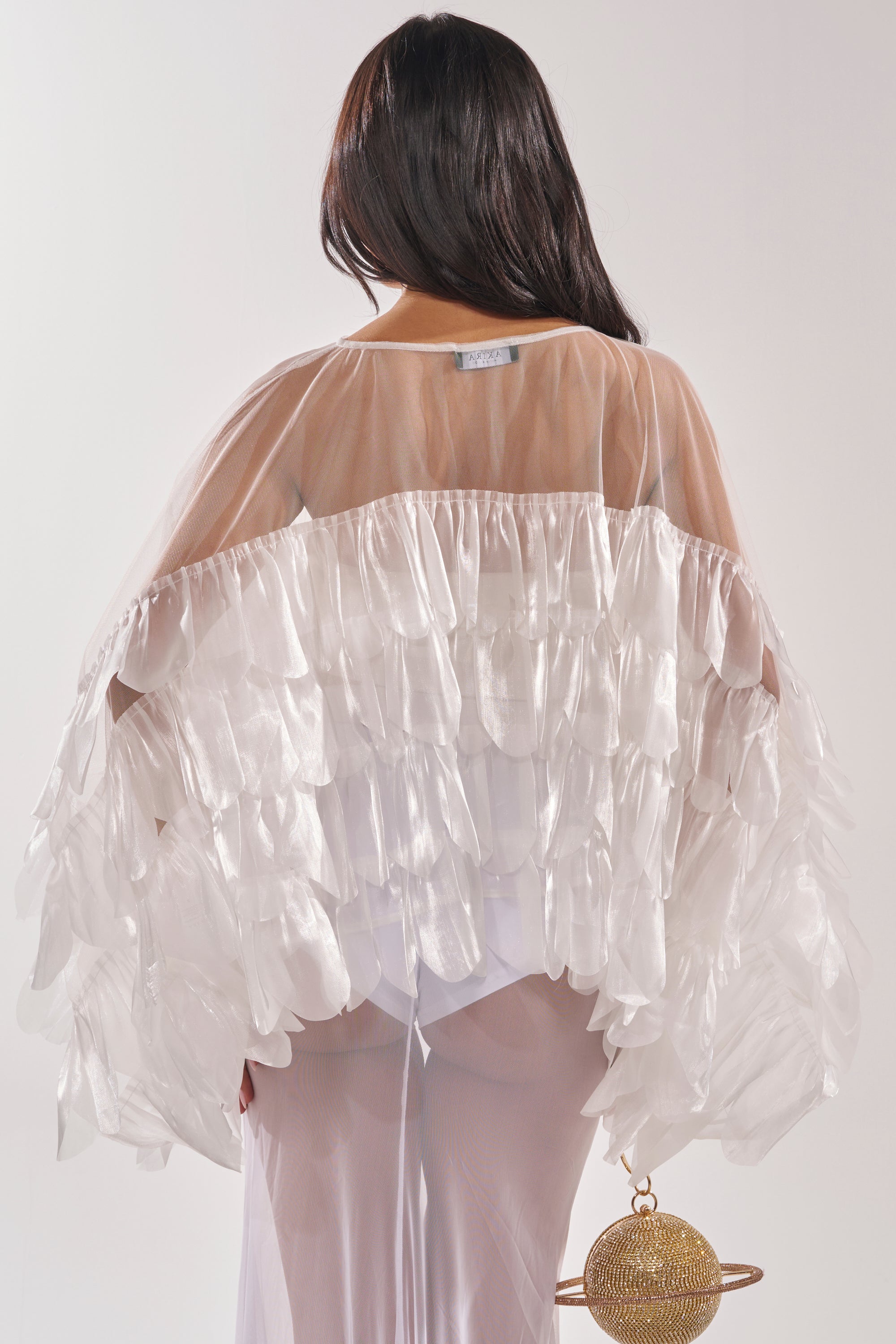 A person with long dark hair is seen from the back, wearing the RUFFLE CITY BLOUSE with sheer mesh panels and a translucent skirt, holding a spherical gold handbag against a plain light background. 4