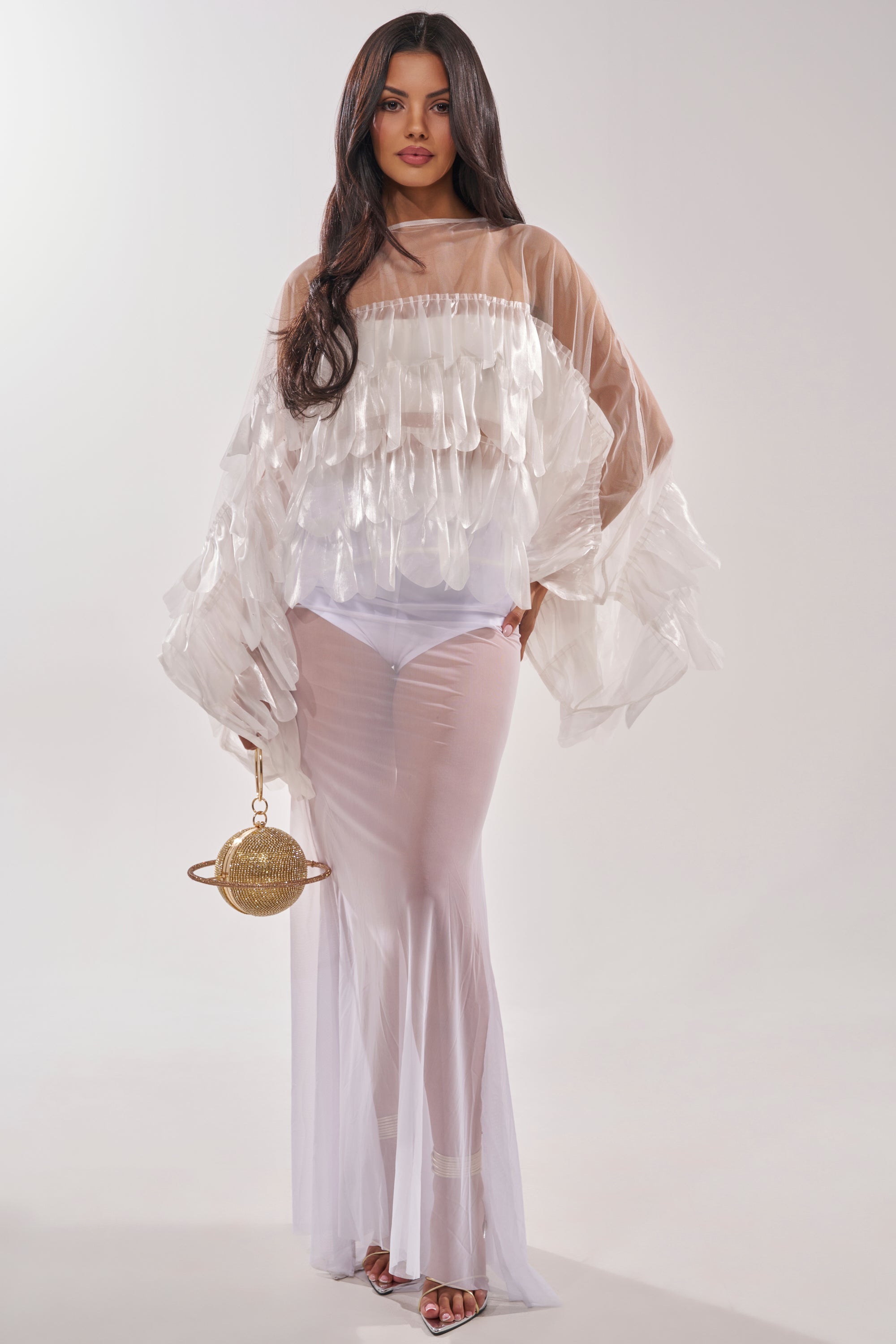 A woman wears the RUFFLE CITY BLOUSE, featuring sheer layered ruffles and wide sleeves, over a white bodysuit. She stands against a plain light background, accessorizing with a gold circular handbag and heeled sandals. 5
