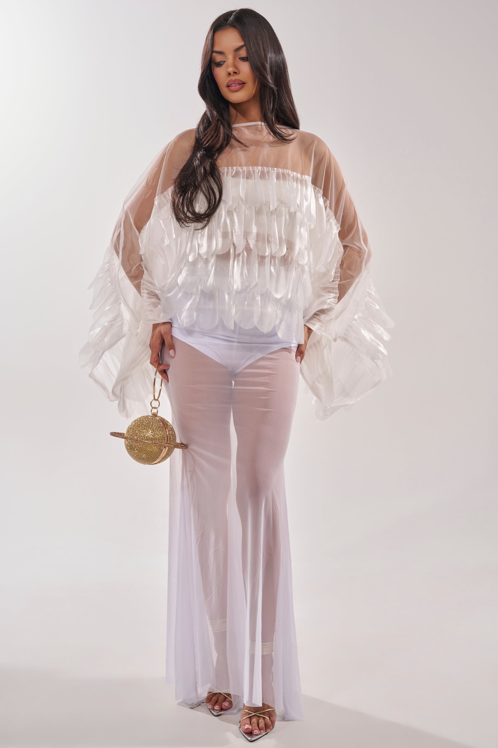 A woman wears the RUFFLE CITY BLOUSE, a sheer white top with layered ruffles, paired with wide, flowing pants. She accessorizes with a small straw bag and strappy gold sandals, her long dark hair styled in loose waves. 6