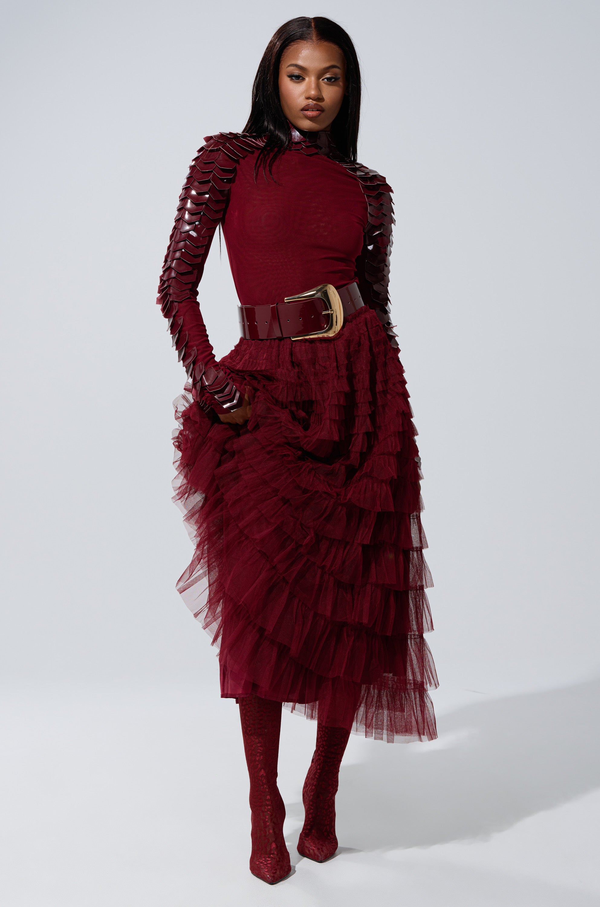 A woman stands confidently against a light gray background, wearing the STARS ALIGNED Mesh Ruffle Maxi Skirt in burgundy with matching boots and a deep red, long-sleeve top for a striking silhouette.