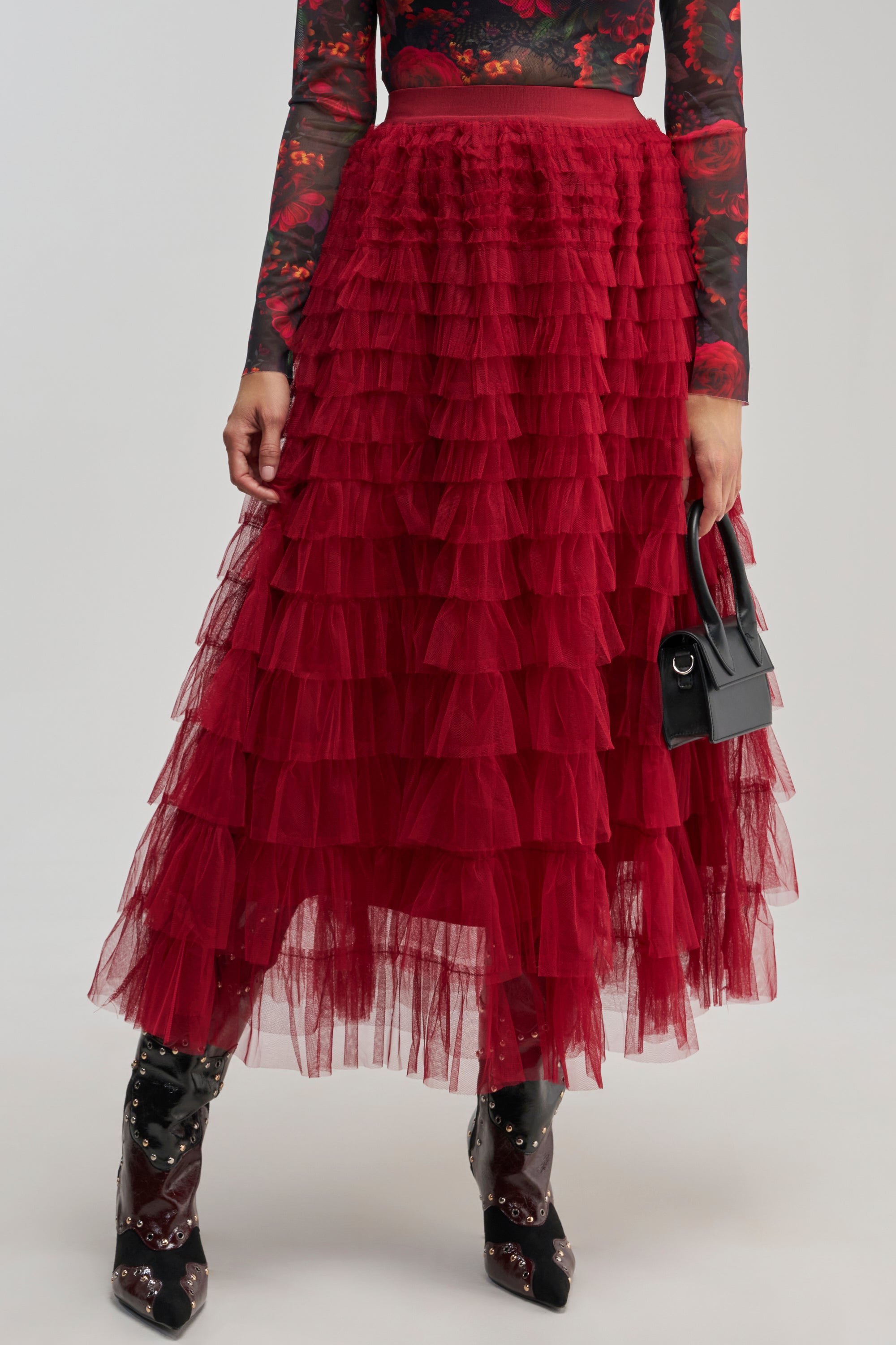 A person wearing the STARS ALIGNED MESH RUFFLE MAXI SKIRT IN BURGUNDY, a long-sleeve floral top, star-printed boots, and holding a small black handbag.