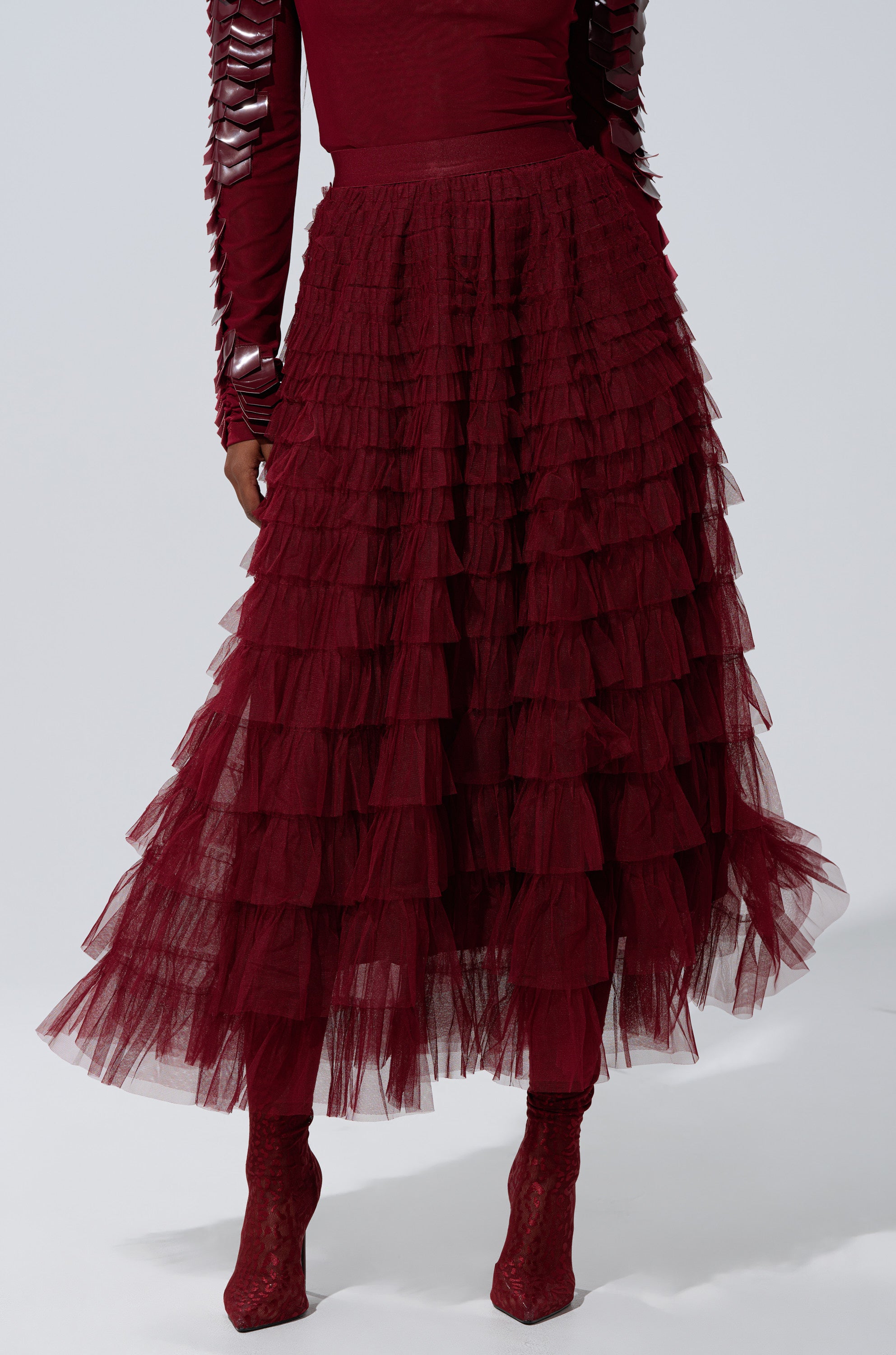 Wearing the STARS ALIGNED MESH RUFFLE MAXI SKIRT IN BURGUNDY, a person pairs it with a matching textured top and burgundy lace-up heeled boots, standing against a plain light background.
