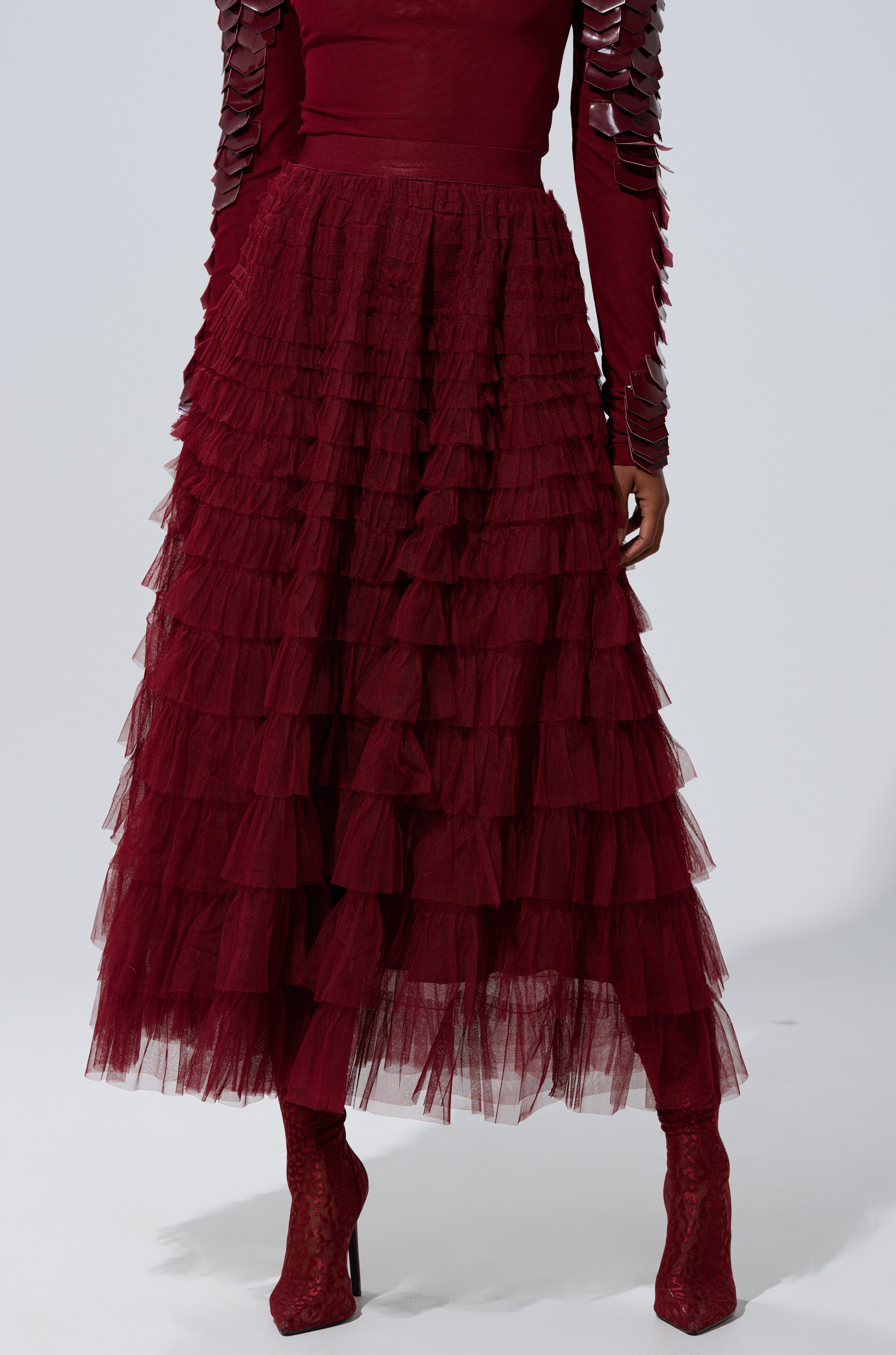 A person wears the STARS ALIGNED MESH RUFFLE MAXI SKIRT IN BURGUNDY with a matching burgundy long-sleeve top and lace heeled boots, standing against a light gray background.