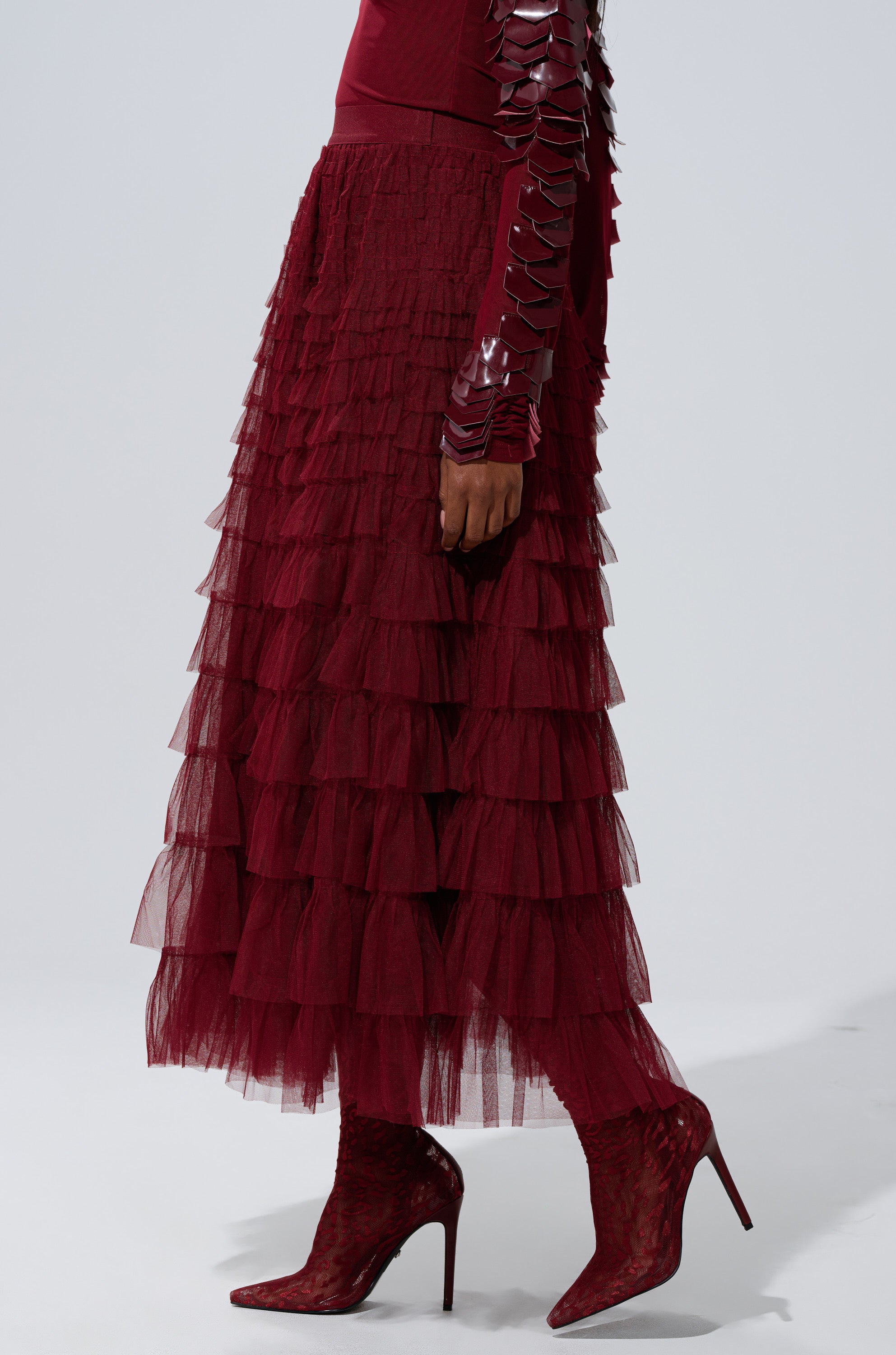 A person wears the STARS ALIGNED MESH RUFFLE MAXI SKIRT IN BURGUNDY with matching pointed-toe high-heeled boots and a long-sleeve top with shiny, segmented arm details, set against a plain light-colored background.