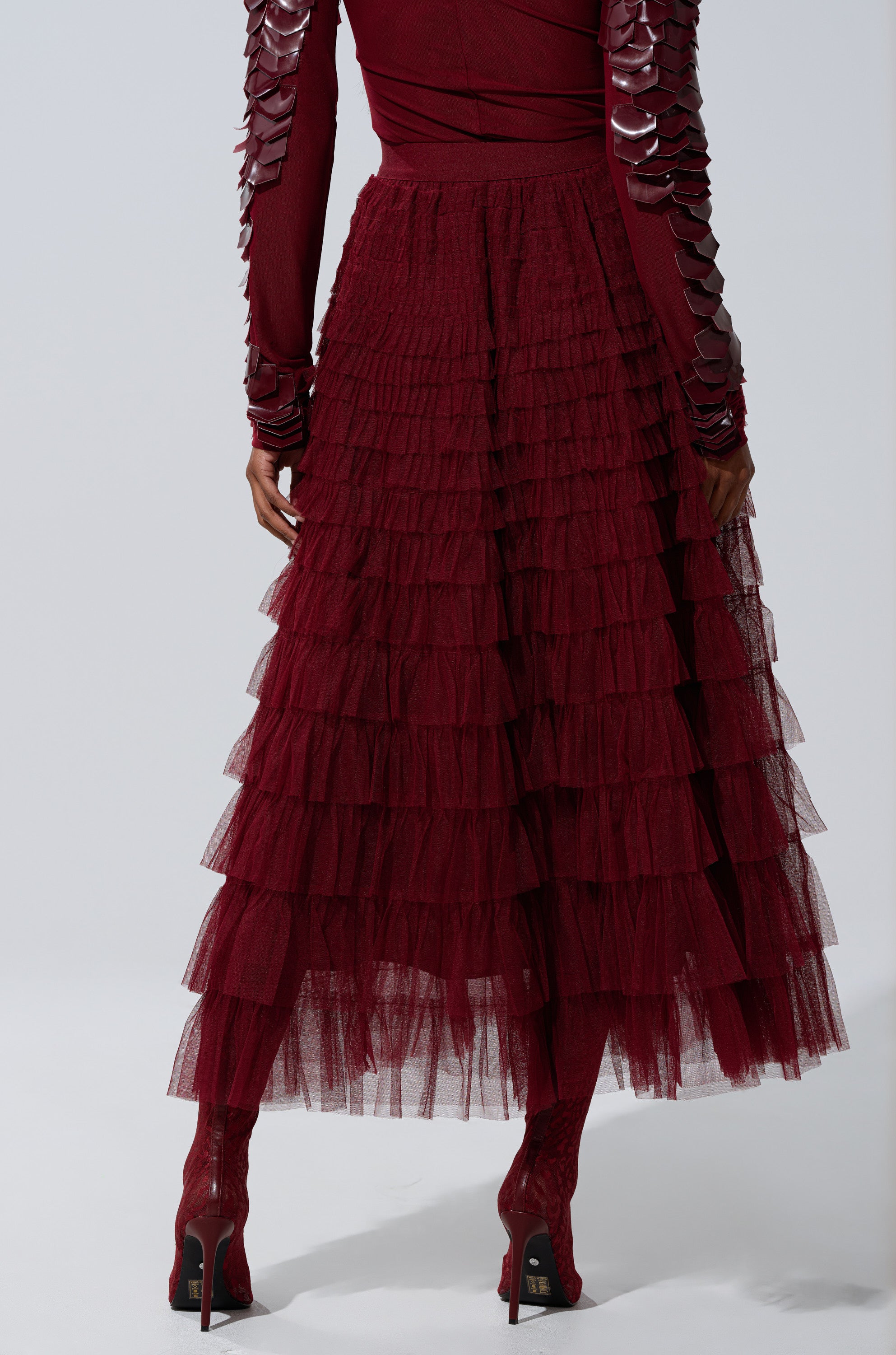 A person is seen from the back against a plain background, wearing the STARS ALIGNED Mesh Ruffle Maxi Skirt in Burgundy, a matching textured top with decorative sleeves, and heeled shoes.