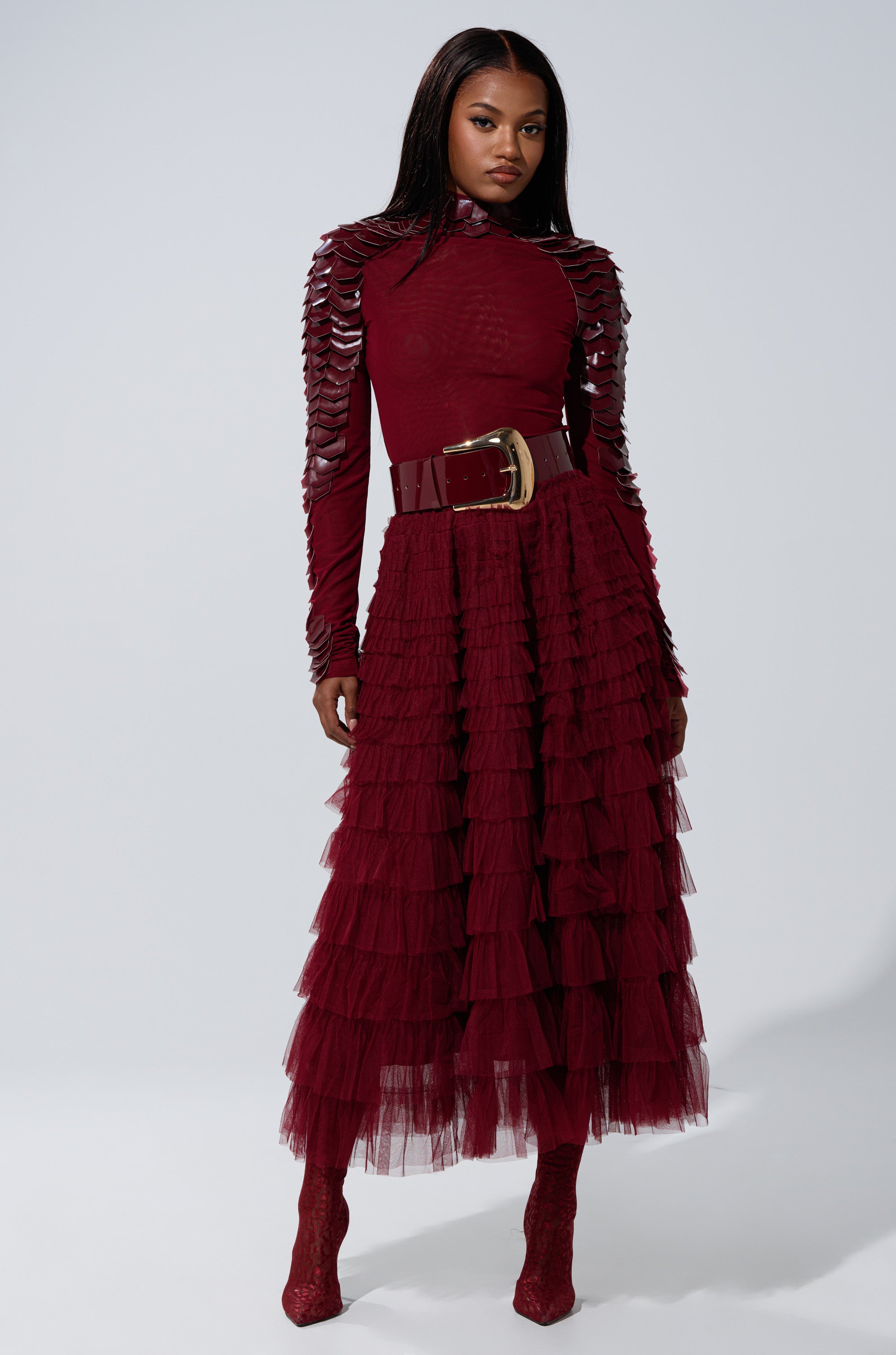 A woman stands confidently in a studio wearing the STARS ALIGNED MESH RUFFLE MAXI SKIRT IN BURGUNDY, styled with layered ruffles, textured sleeves, a wide belt with a gold buckle, and matching red boots.