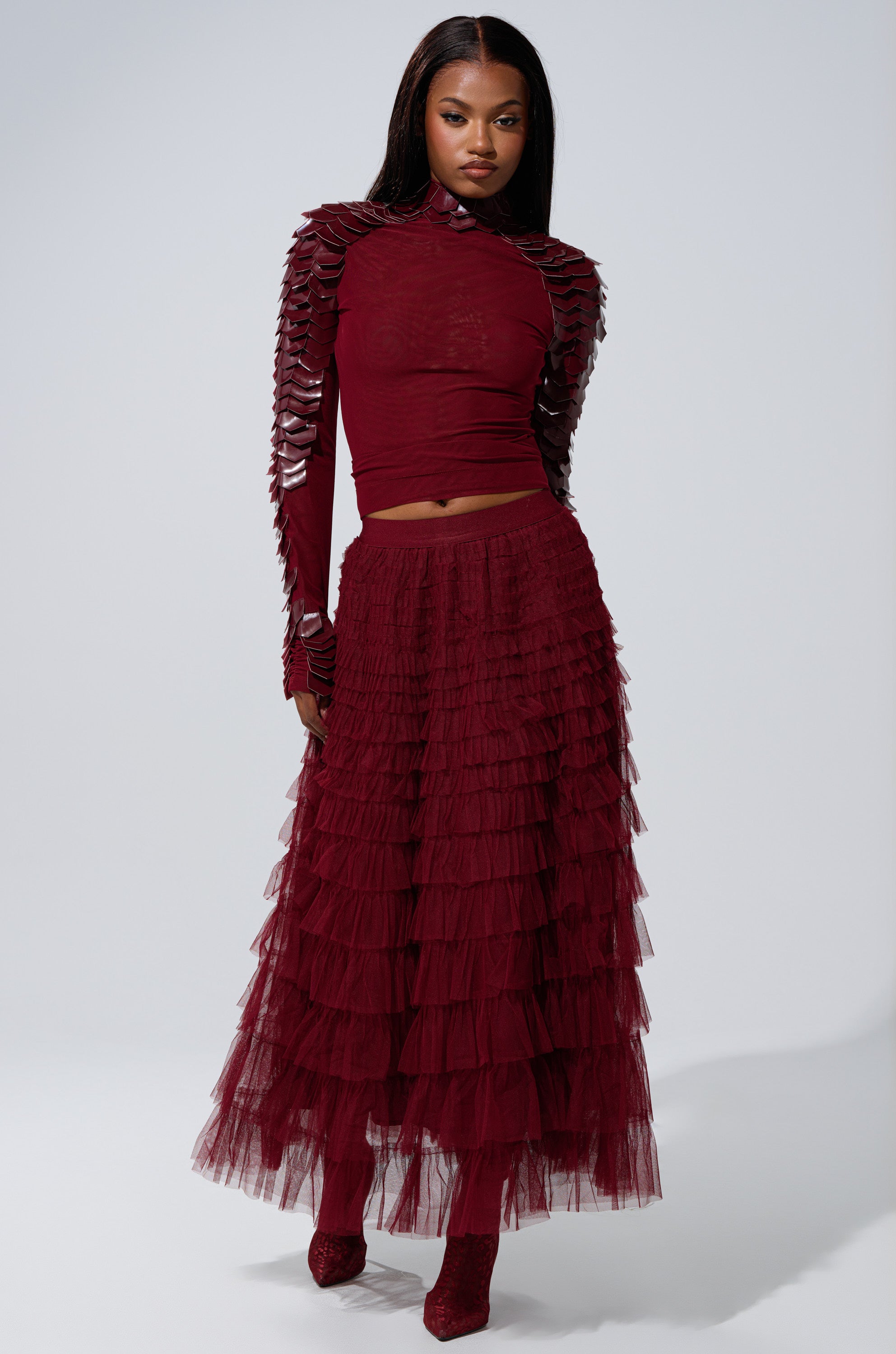 A woman stands against a plain light background wearing the STARS ALIGNED MESH RUFFLE MAXI SKIRT IN BURGUNDY, paired with a dark red, long-sleeved top featuring layered sleeve details and red shoes.