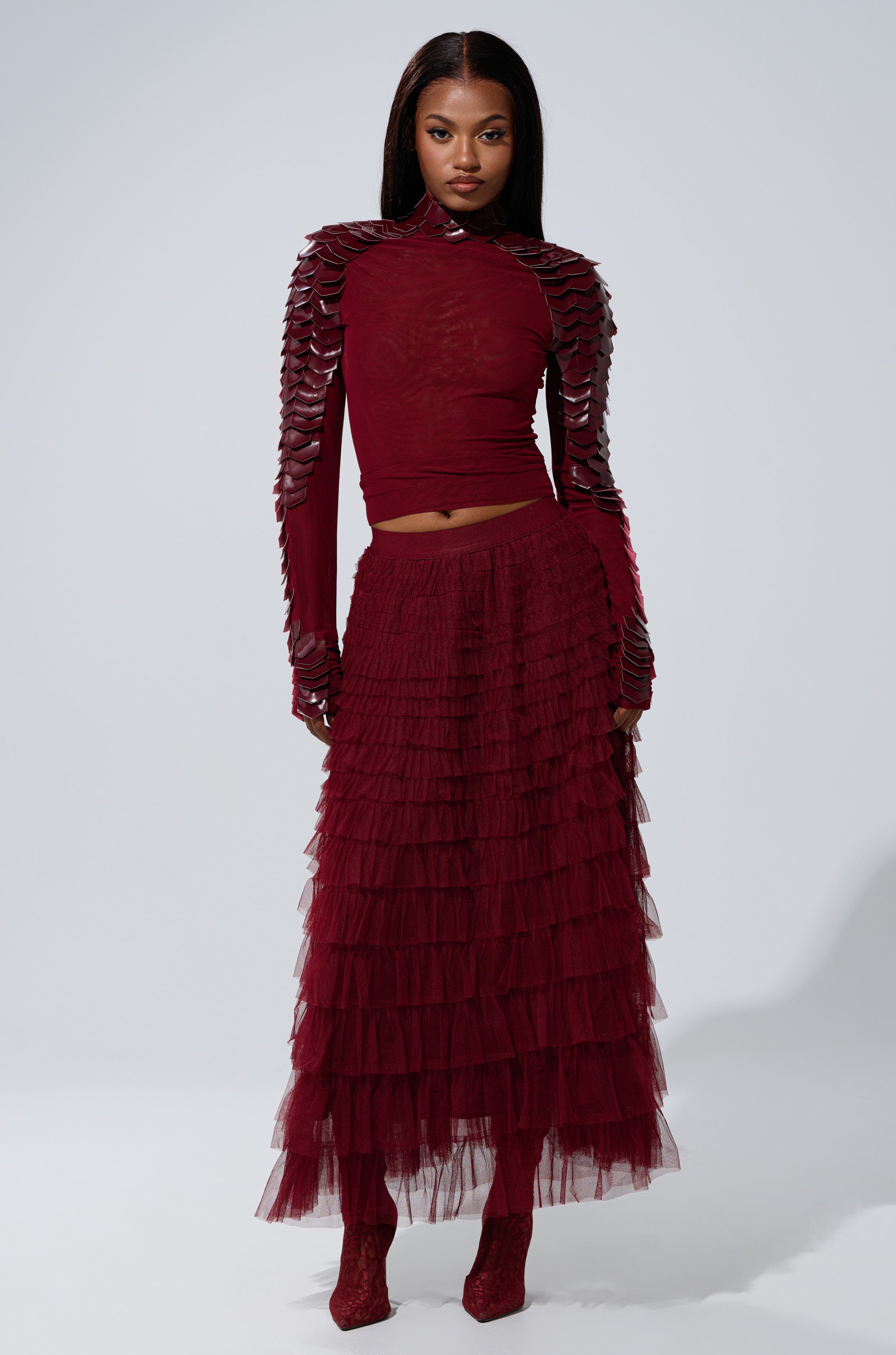 A woman stands against a plain light background wearing the STARS ALIGNED MESH RUFFLE MAXI SKIRT IN BURGUNDY, featuring textured, layered details, paired with a matching top with sculpted sleeves.