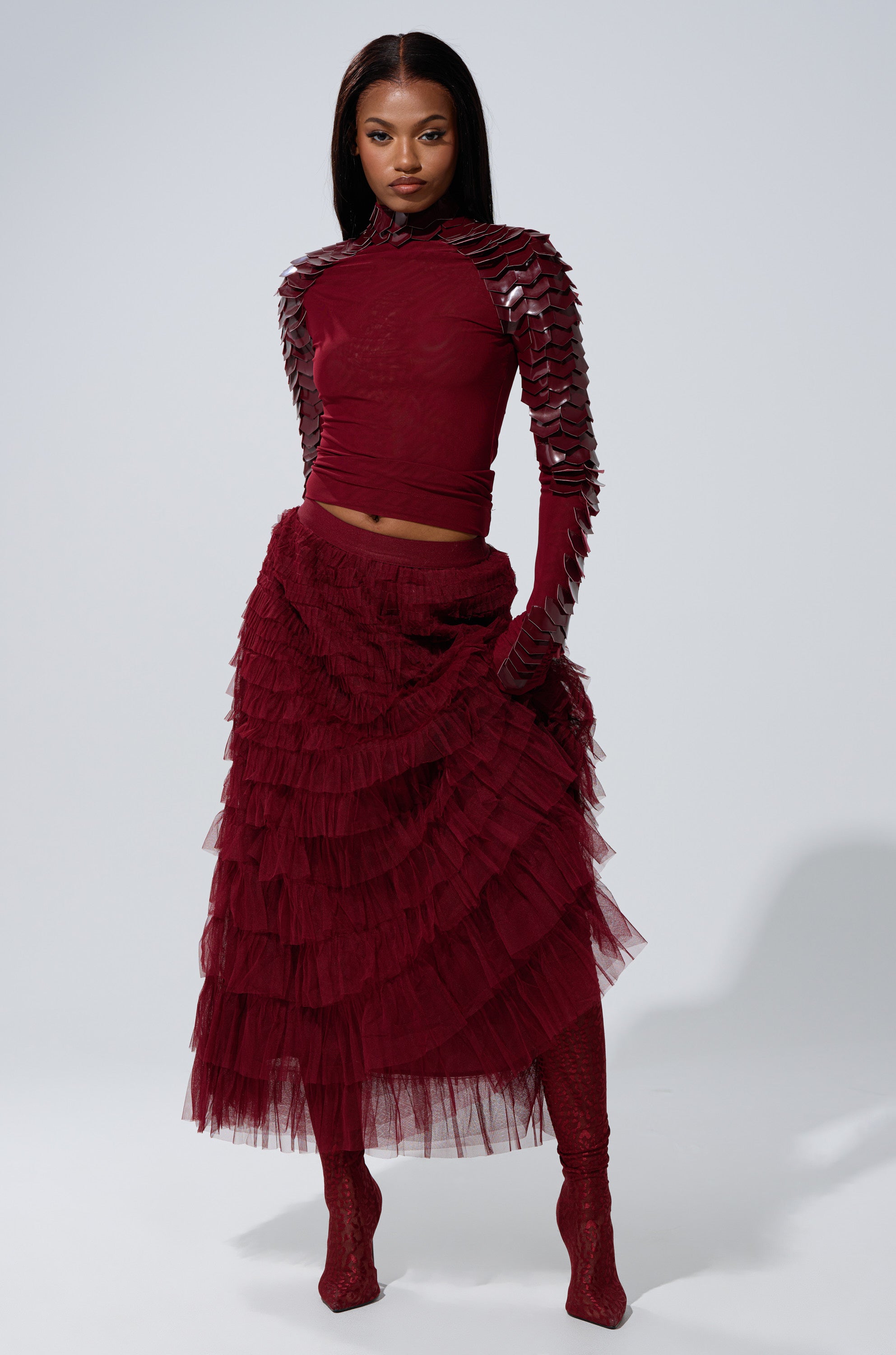 A woman stands confidently in the STARS ALIGNED MESH RUFFLE MAXI SKIRT IN BURGUNDY, paired with a textured top, red tights, and heeled boots against a plain light background.