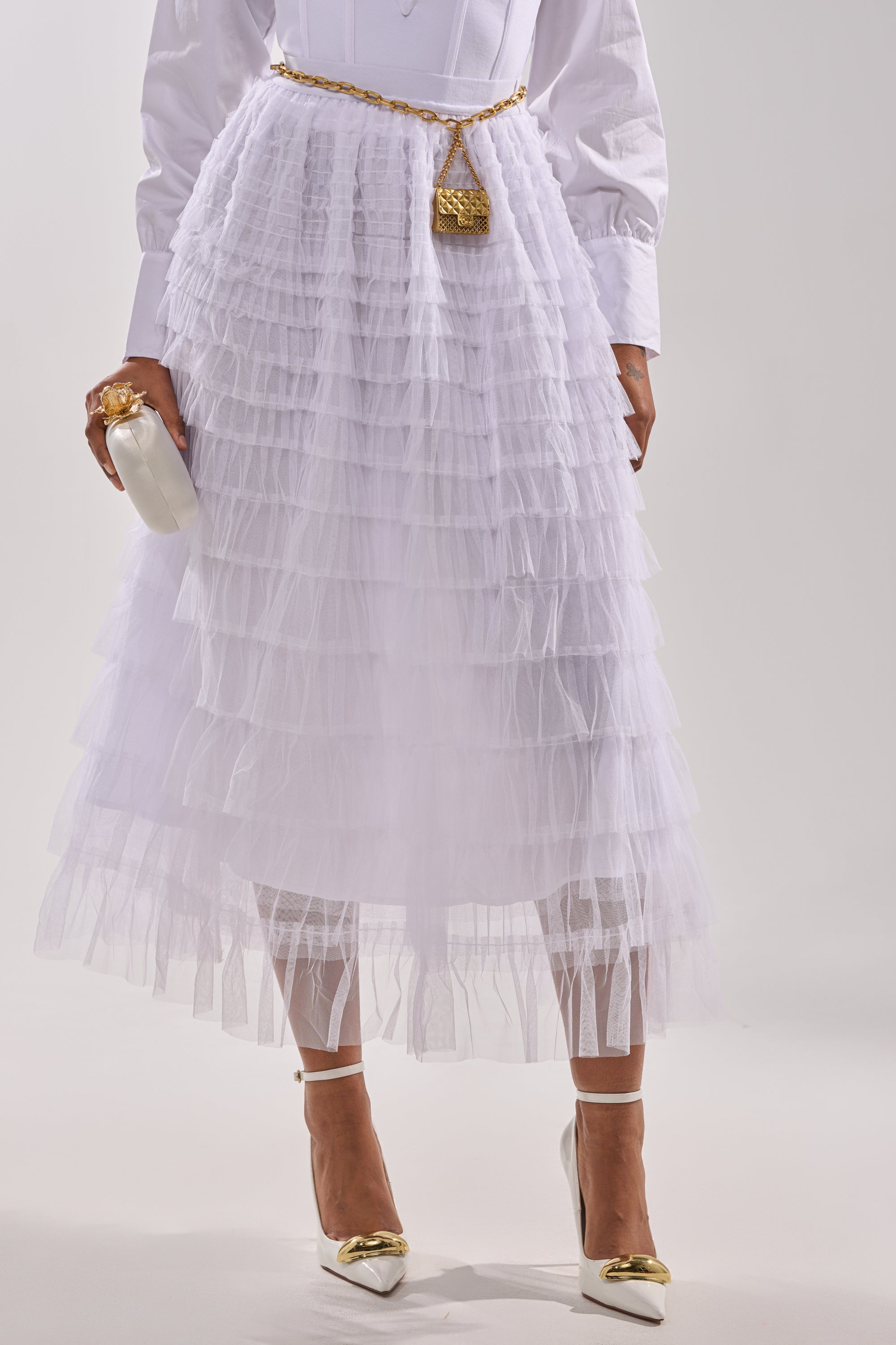 A woman wears the STARS ALIGNED MESH RUFFLE MAXI SKIRT IN WHITE paired with a white long-sleeved top, white heels with gold accents, a thin gold belt bag, and holds a white clutch with a gold clasp against a plain light background. 0