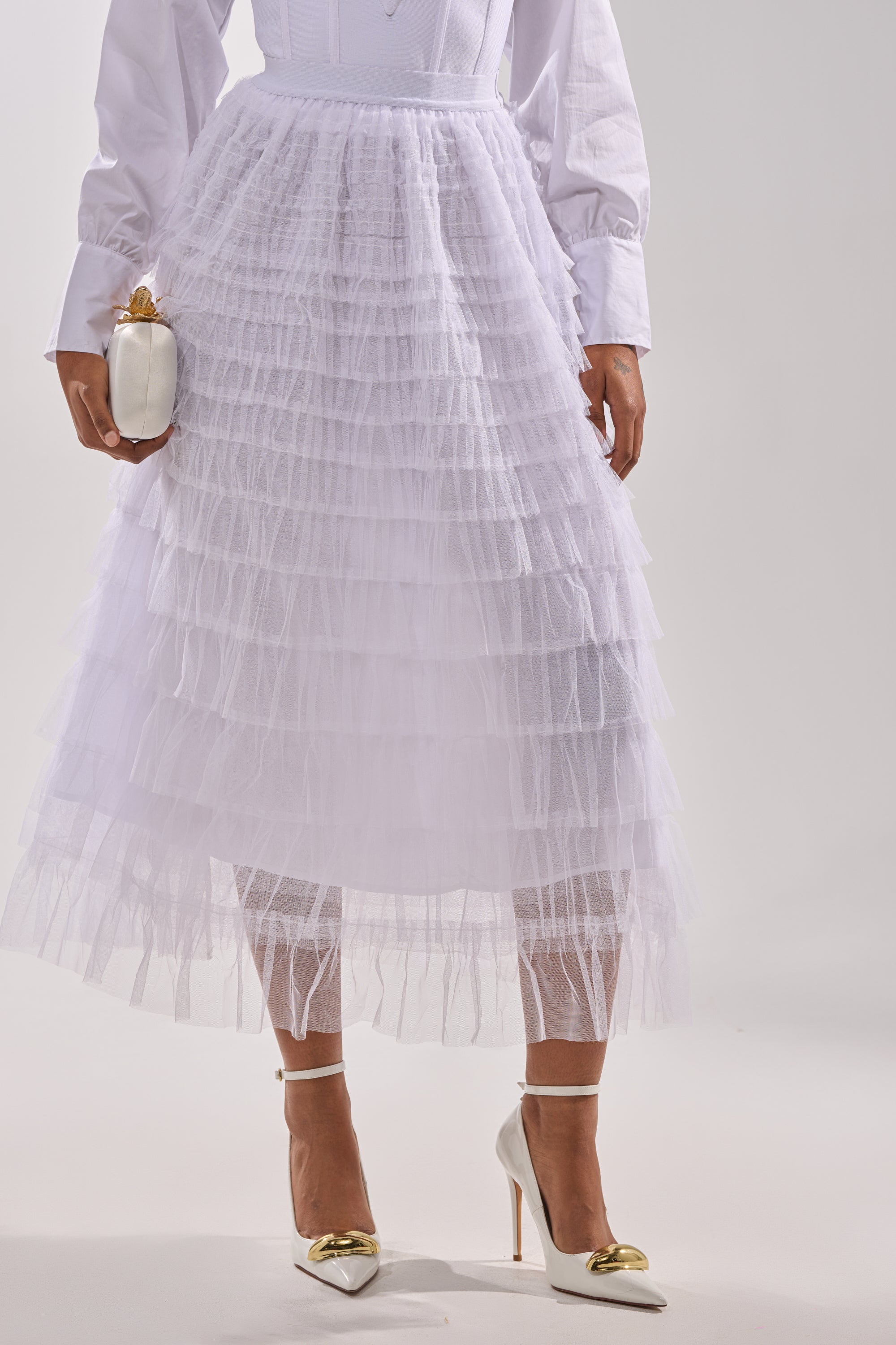 Wearing the STARS ALIGNED MESH RUFFLE MAXI SKIRT IN WHITE, a white button-down top, white high heels with gold detail, and holding a white clutch with a gold clasp, this person stands against a plain background. 1