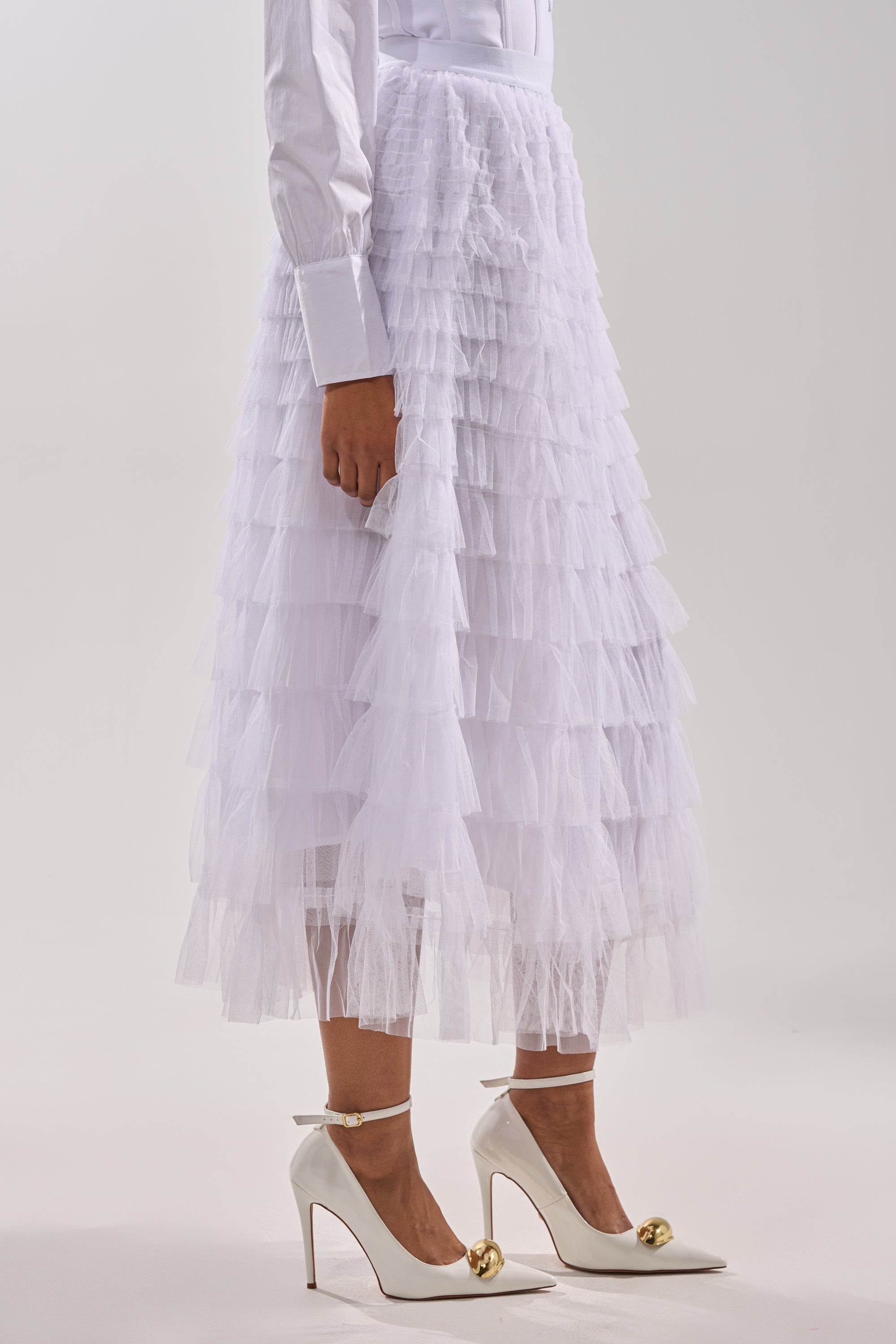 A person stands against a light background wearing the STARS ALIGNED MESH RUFFLE MAXI SKIRT IN WHITE, white high heels with gold ball details, and a white long-sleeve shirt with wide cuffs. 2