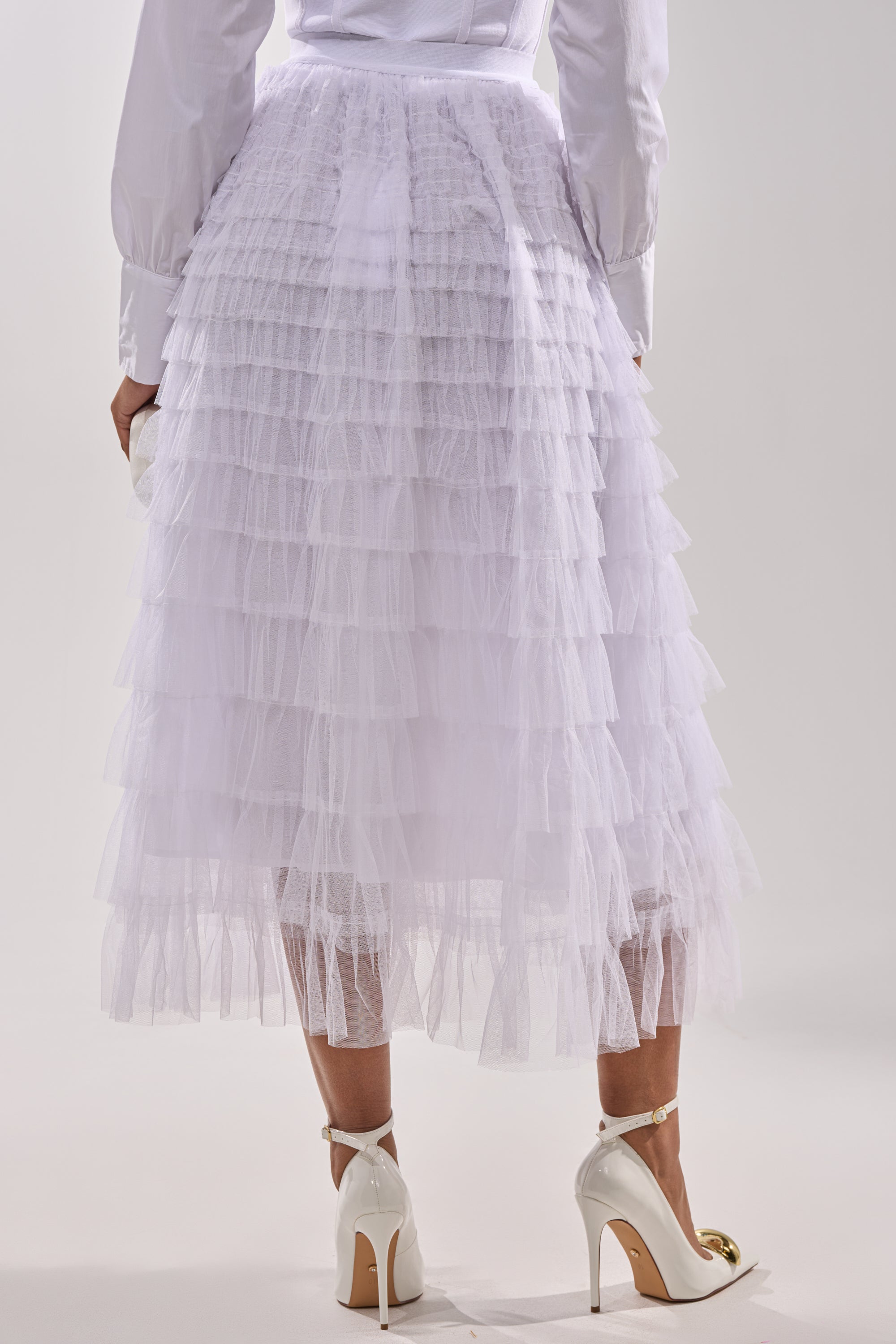 Wearing the STARS ALIGNED MESH RUFFLE MAXI SKIRT IN WHITE with a long-sleeve blouse and high heels, a person stands against a light background, viewed from behind. 3