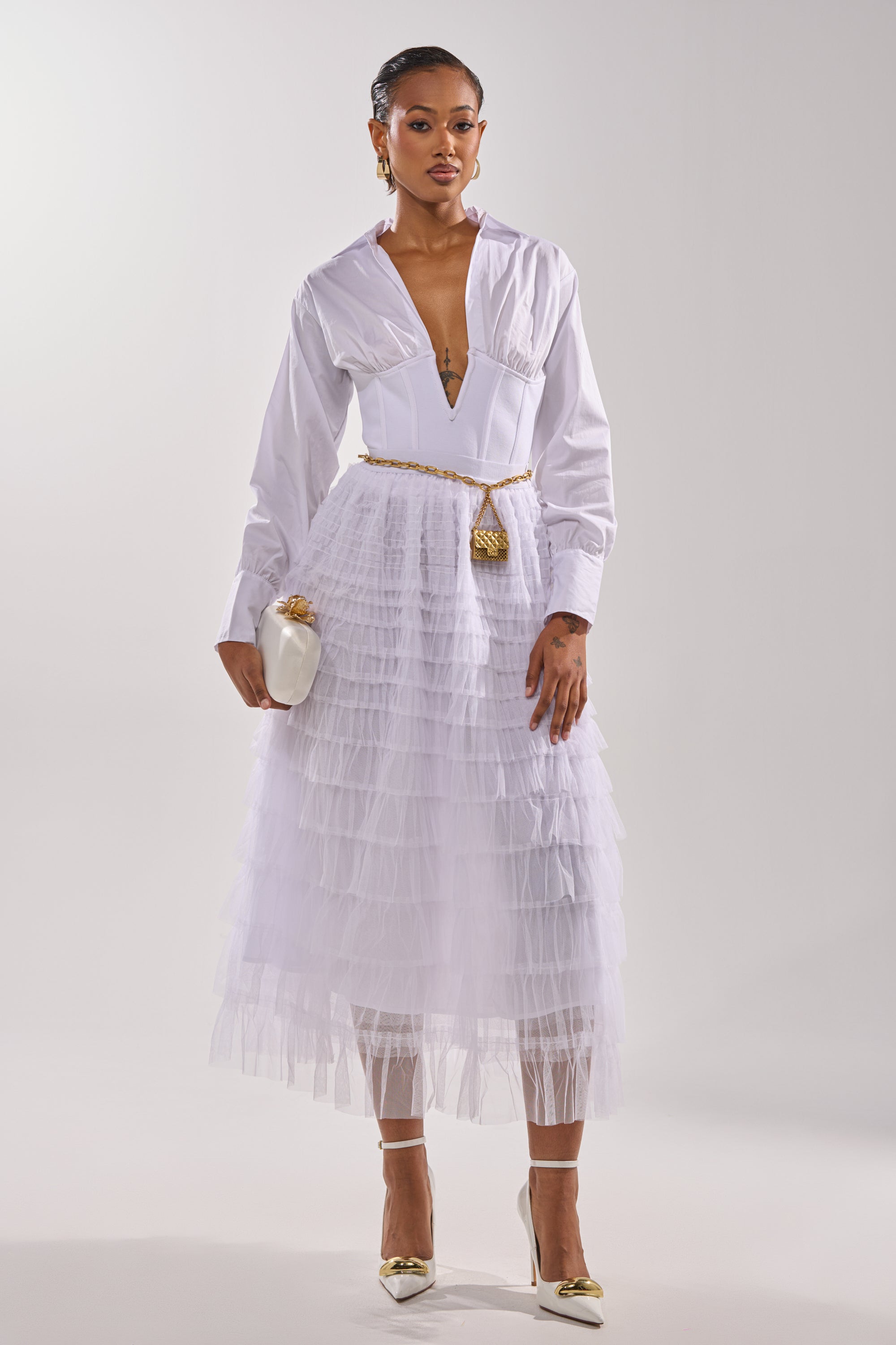 A stylish woman wears the STARS ALIGNED MESH RUFFLE MAXI SKIRT IN WHITE with a deep V blouse, gold belt, white clutch, gold earrings, and heels, standing against a plain light background. 4