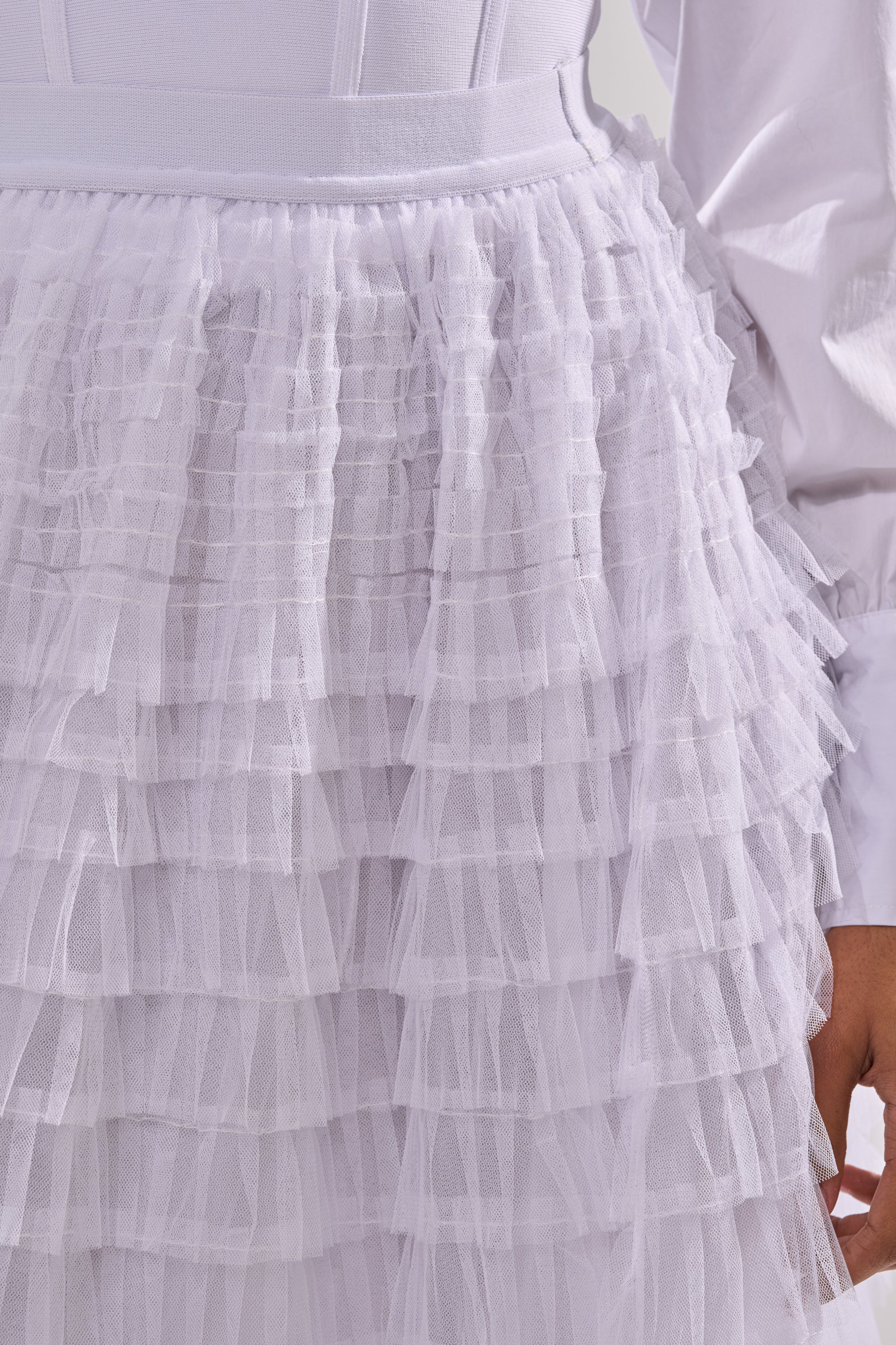 Close-up of a person wearing the STARS ALIGNED MESH RUFFLE MAXI SKIRT IN WHITE, featuring multiple layers of ruffled tulle. Their right arm, dressed in a long white sleeve, is partly visible. 5