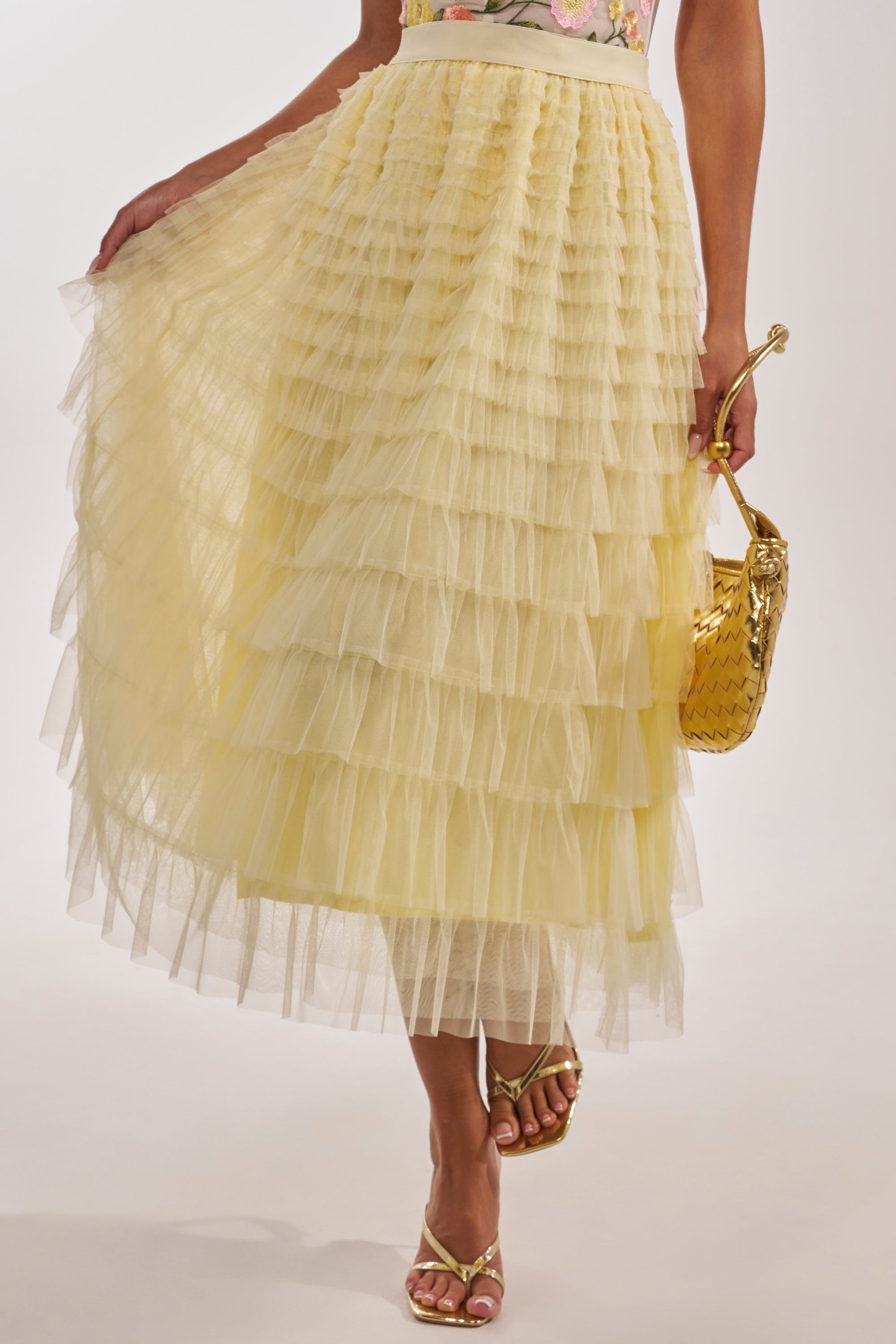 A woman’s lower half is shown wearing the STARS ALIGNED MESH RUFFLE MAXI SKIRT IN BABY YELLOW, featuring tiered ruffles, paired with gold strappy heels and a gold textured handbag. The skirt’s voluminous layers create a standout look. 1