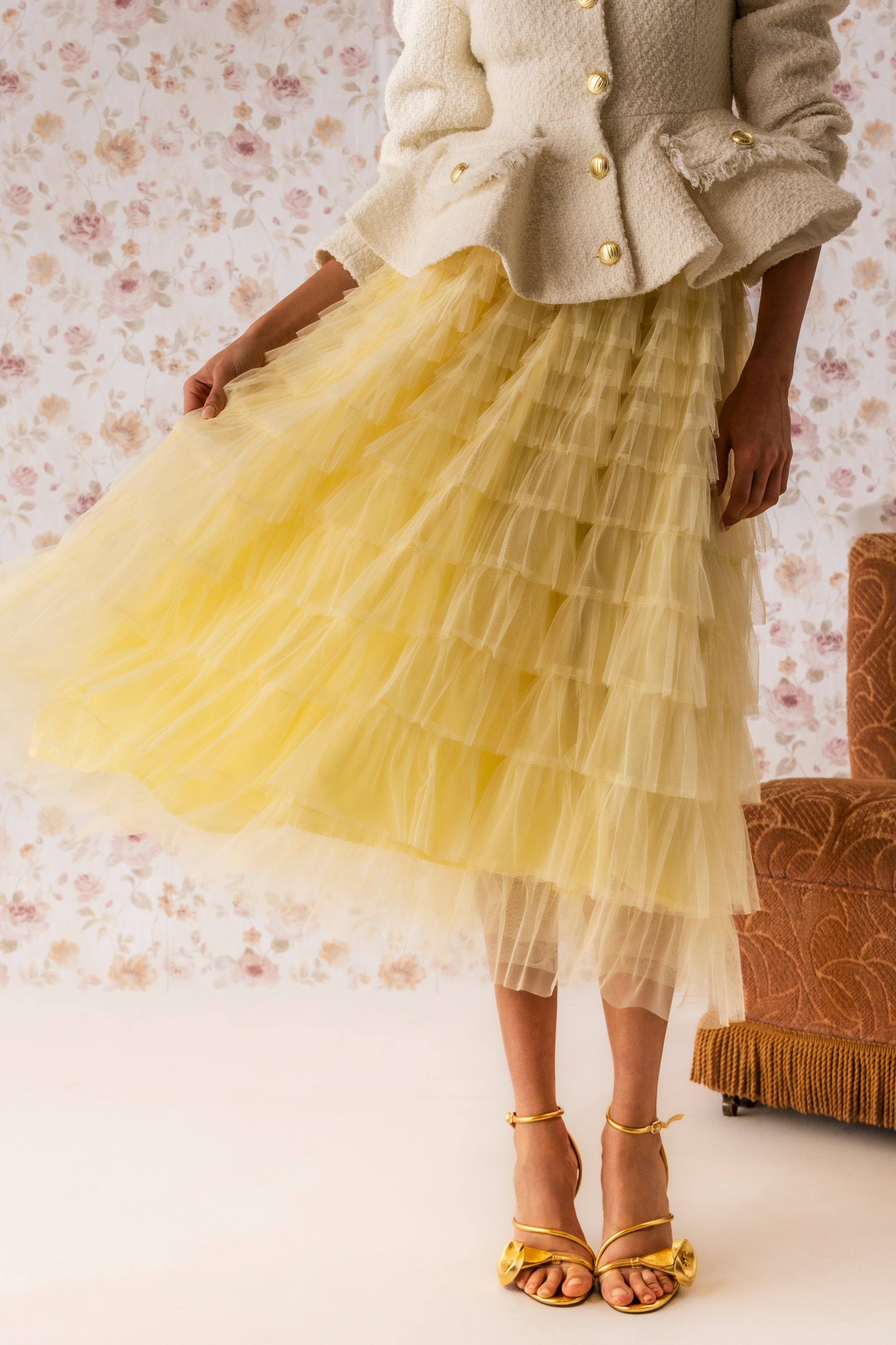 Wearing a textured cream jacket with gold buttons and the STARS ALIGNED MESH RUFFLE MAXI SKIRT IN BABY YELLOW, paired with yellow strappy heels, a person stands before floral wallpaper beside a brown upholstered chair. 0