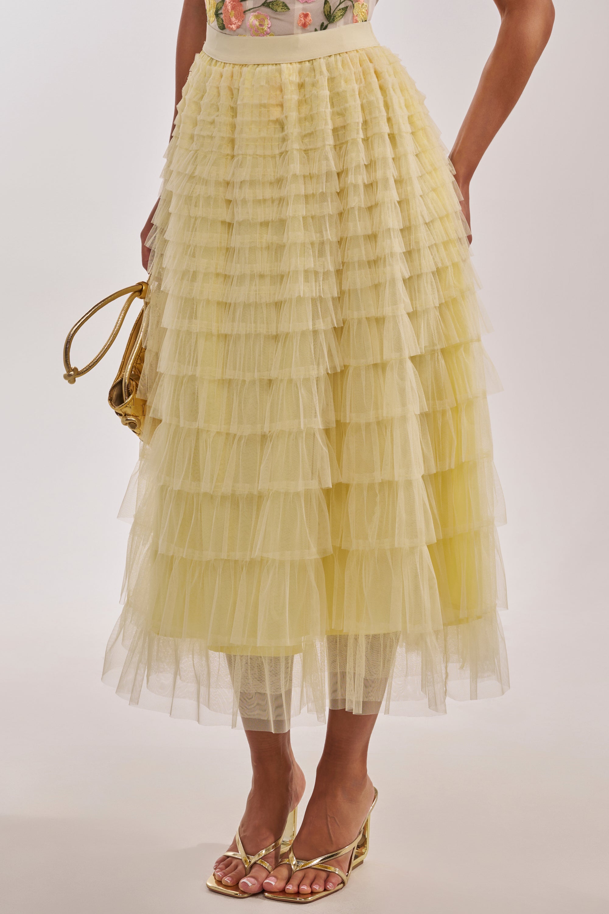 A person wears the STARS ALIGNED MESH RUFFLE MAXI SKIRT IN BABY YELLOW, paired with a floral top, gold strappy heels, and a small gold handbag. The image focuses on the outfit from waist to feet. 2