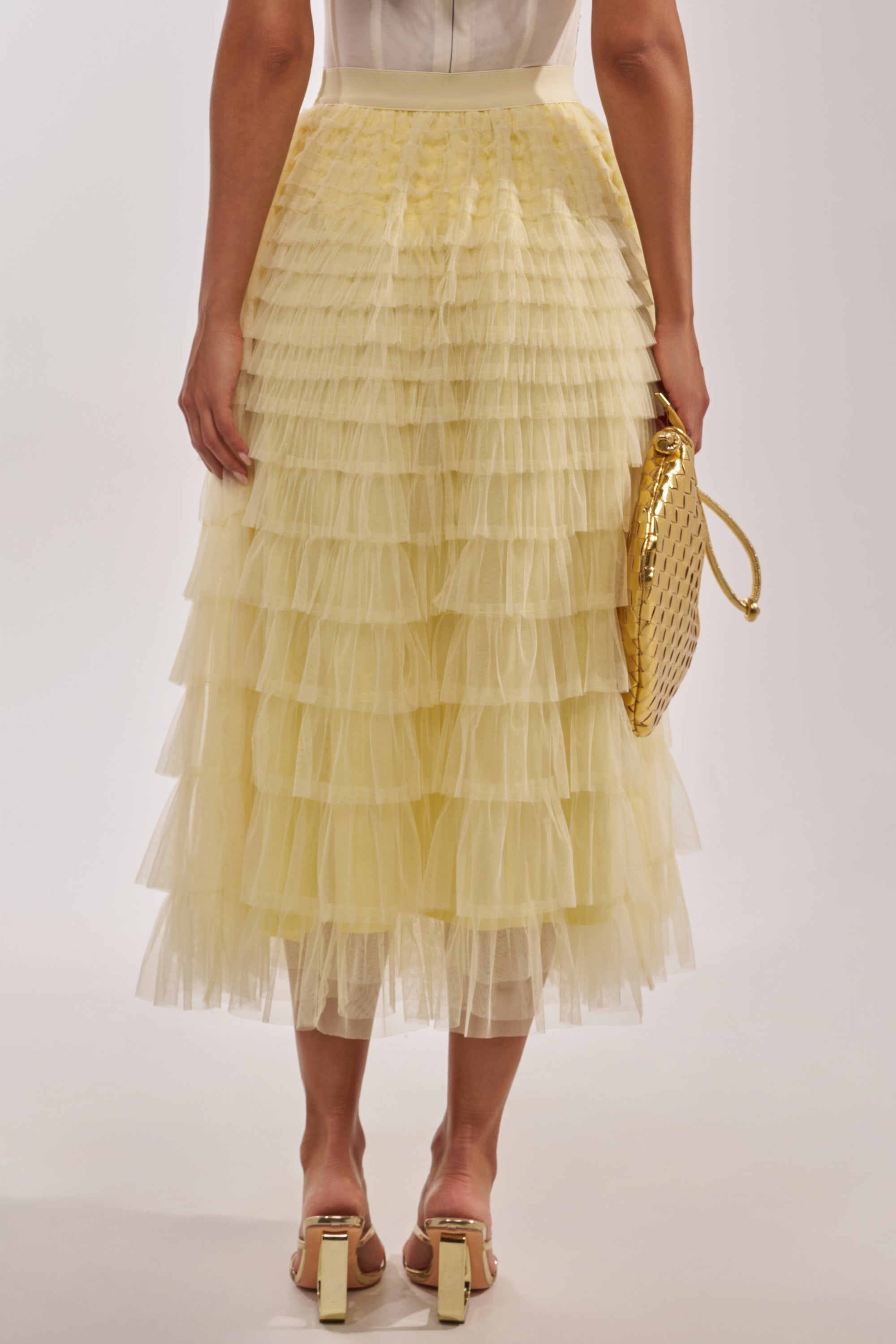 A woman in the STARS ALIGNED MESH RUFFLE MAXI SKIRT IN BABY YELLOW, paired with light heeled sandals and a gold woven handbag, is photographed from behind to showcase the skirt’s delicate, tiered mesh ruffles. 4