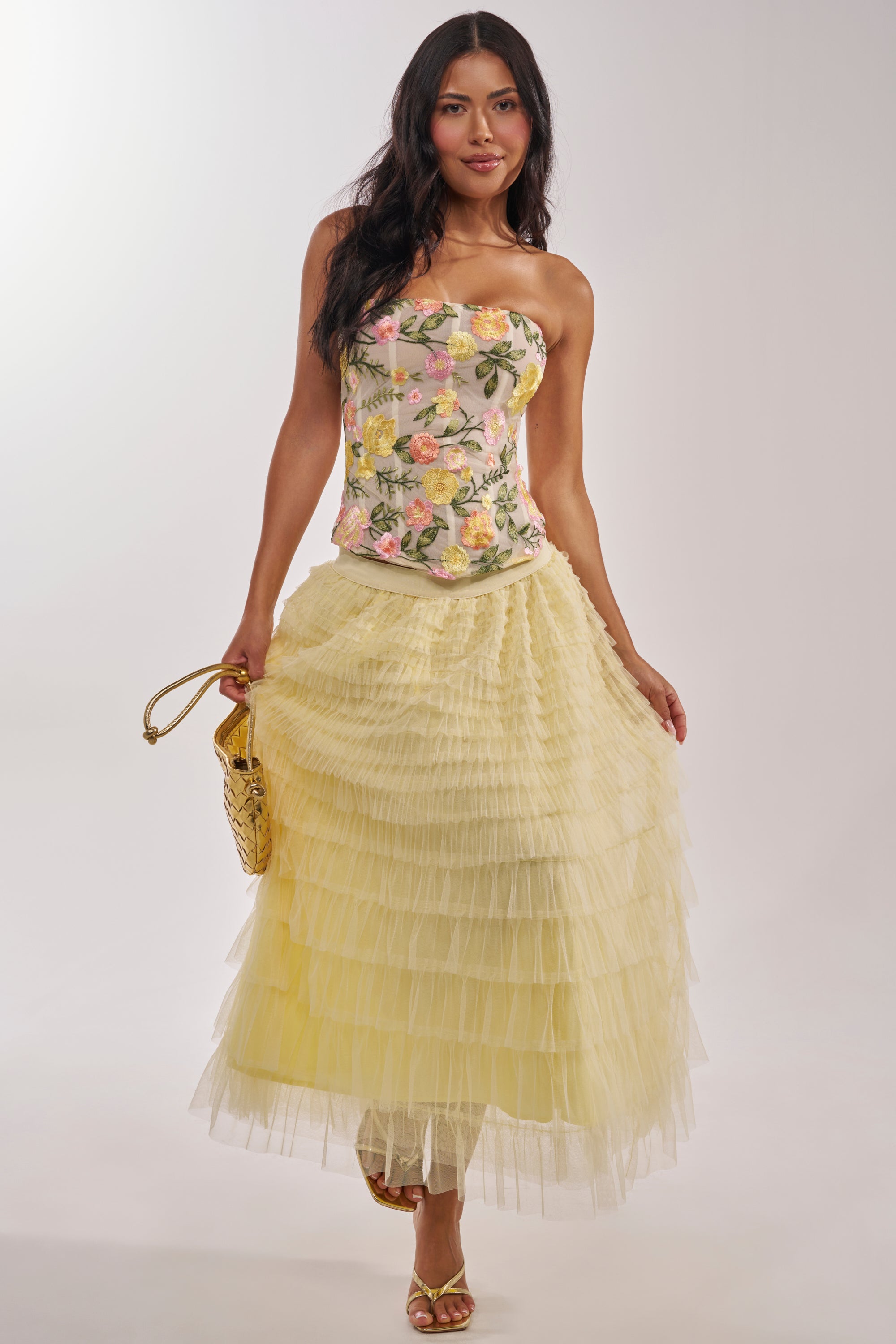 A woman in a strapless floral top and the STARS ALIGNED MESH RUFFLE MAXI SKIRT IN BABY YELLOW holds a gold clutch, wears gold heels, and stands against a plain light background, smiling softly with long dark hair. 5