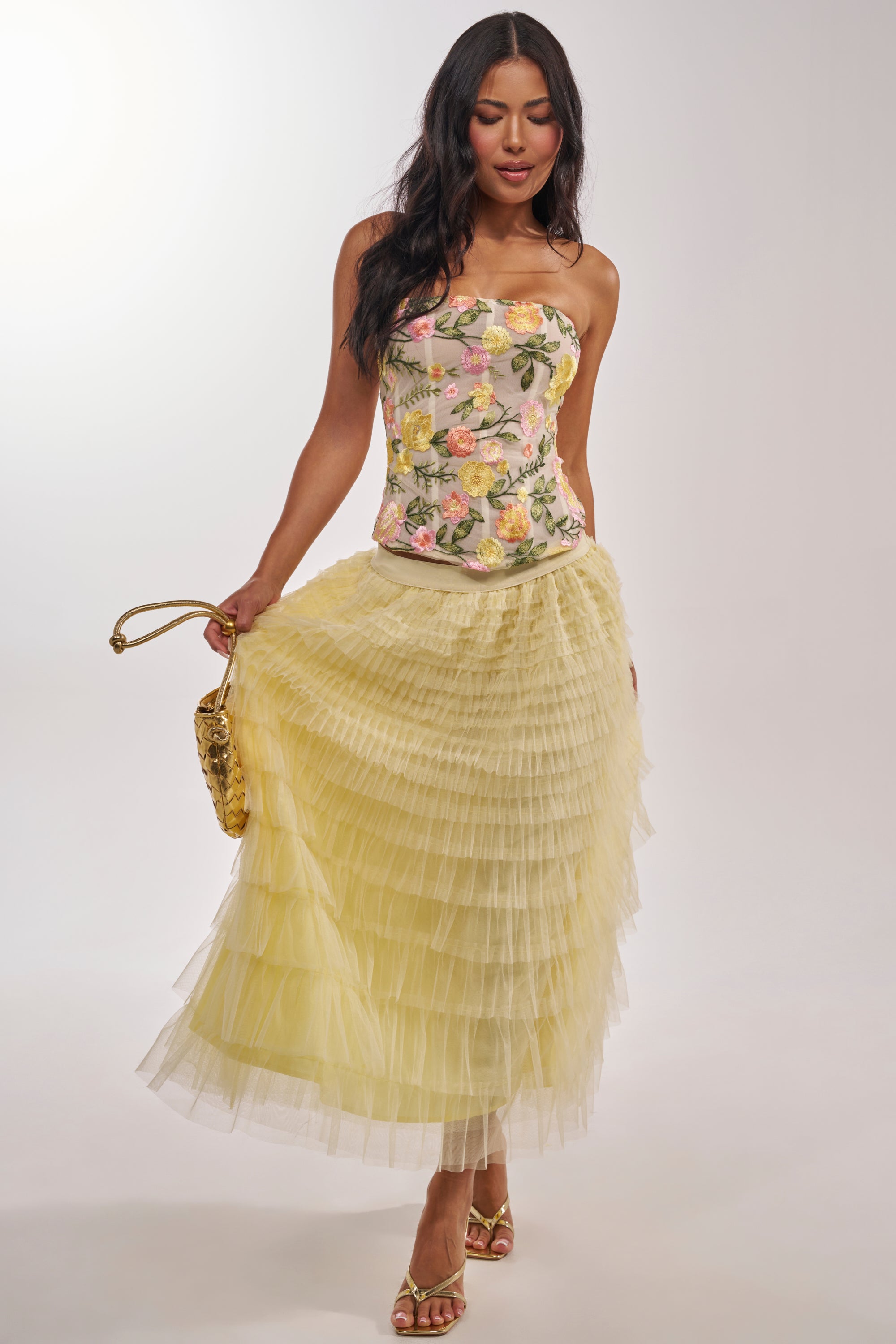 Woman wearing the STARS ALIGNED MESH RUFFLE MAXI SKIRT IN BABY YELLOW with a floral strapless top, gold purse, and gold heeled sandals, standing against a plain light background. 6