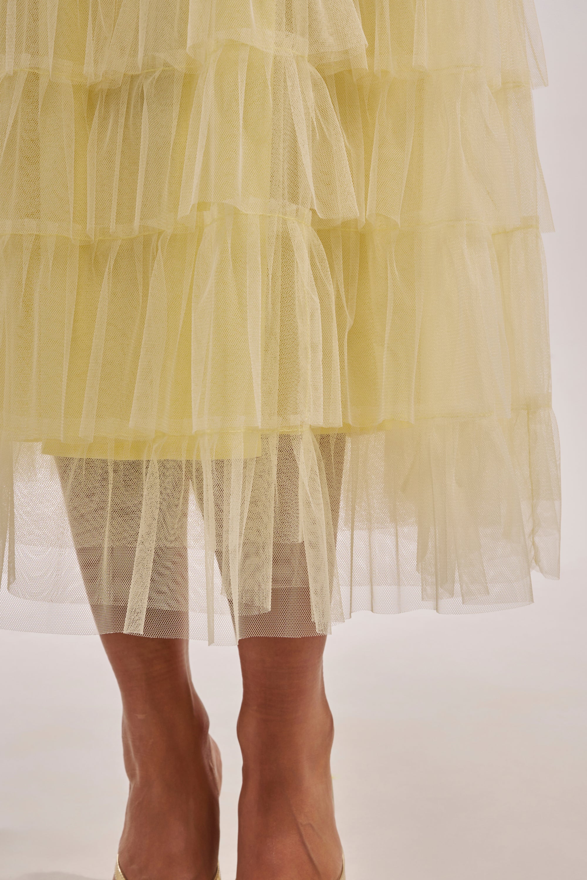 A close-up of someone wearing the STARS ALIGNED MESH RUFFLE MAXI SKIRT IN BABY YELLOW, featuring sheer, tiered layers for a textured look with lower legs and feet visible beneath the ruffled hem. 8